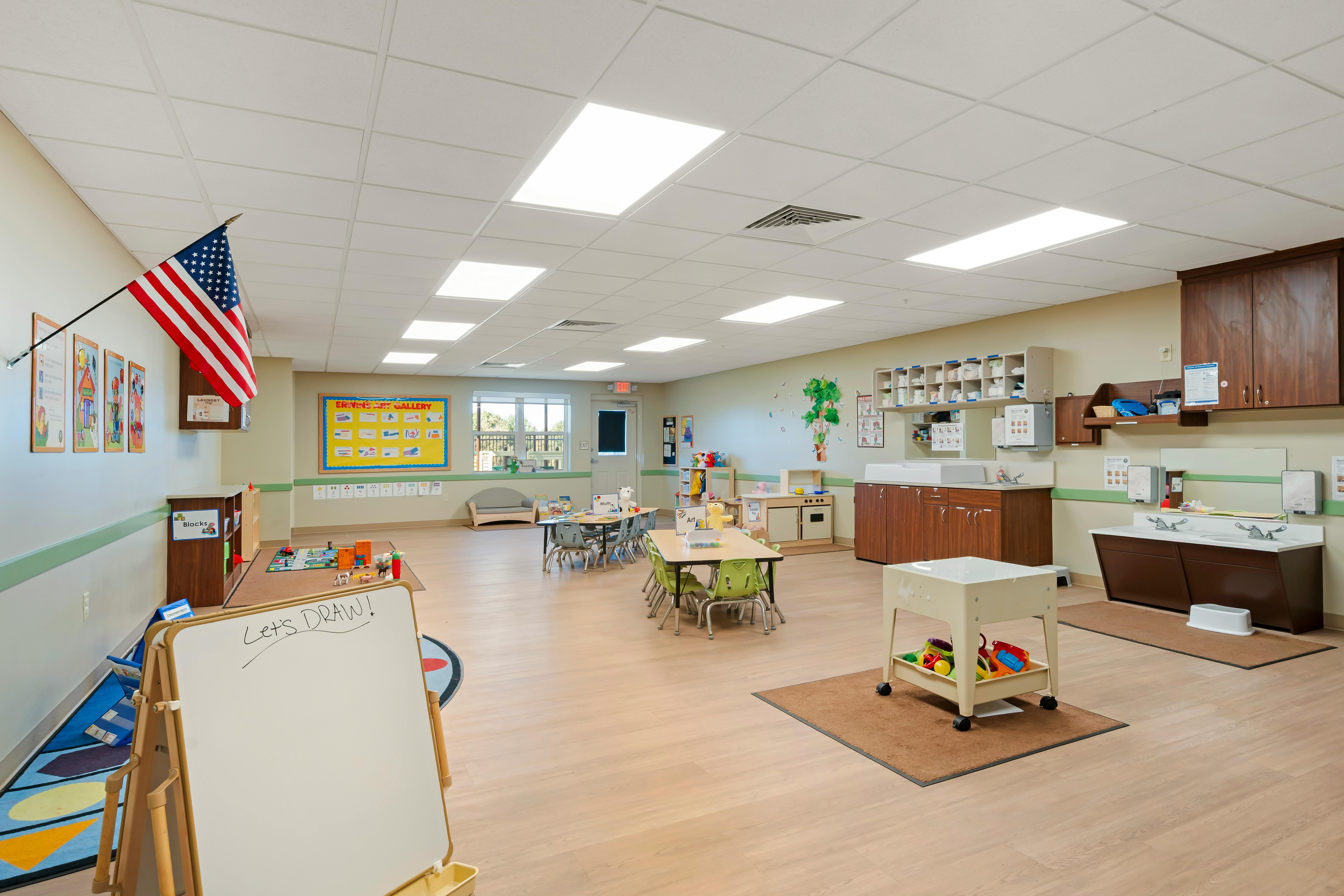 Classroom with tables, chairs, toys, whiteboard with "Let's Draw!" and sinks