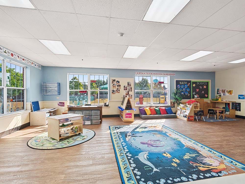 Classroom with large windows, bright lighting, colorful rugs, and various learning stations