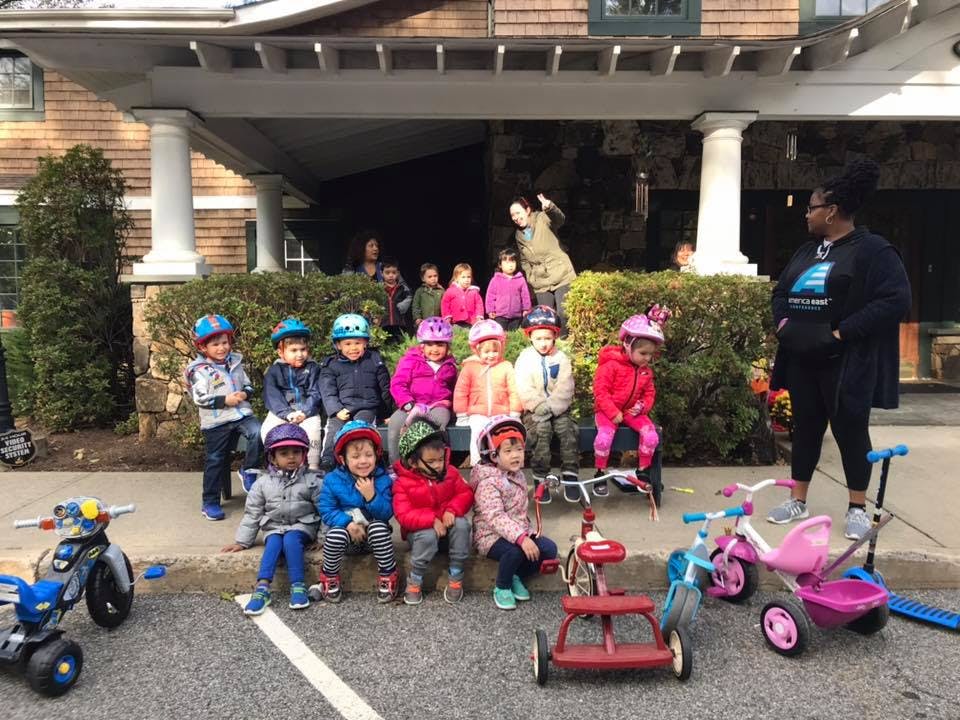 Children wearing helmets sitting on a curb with tricycles and an adult standing nearby