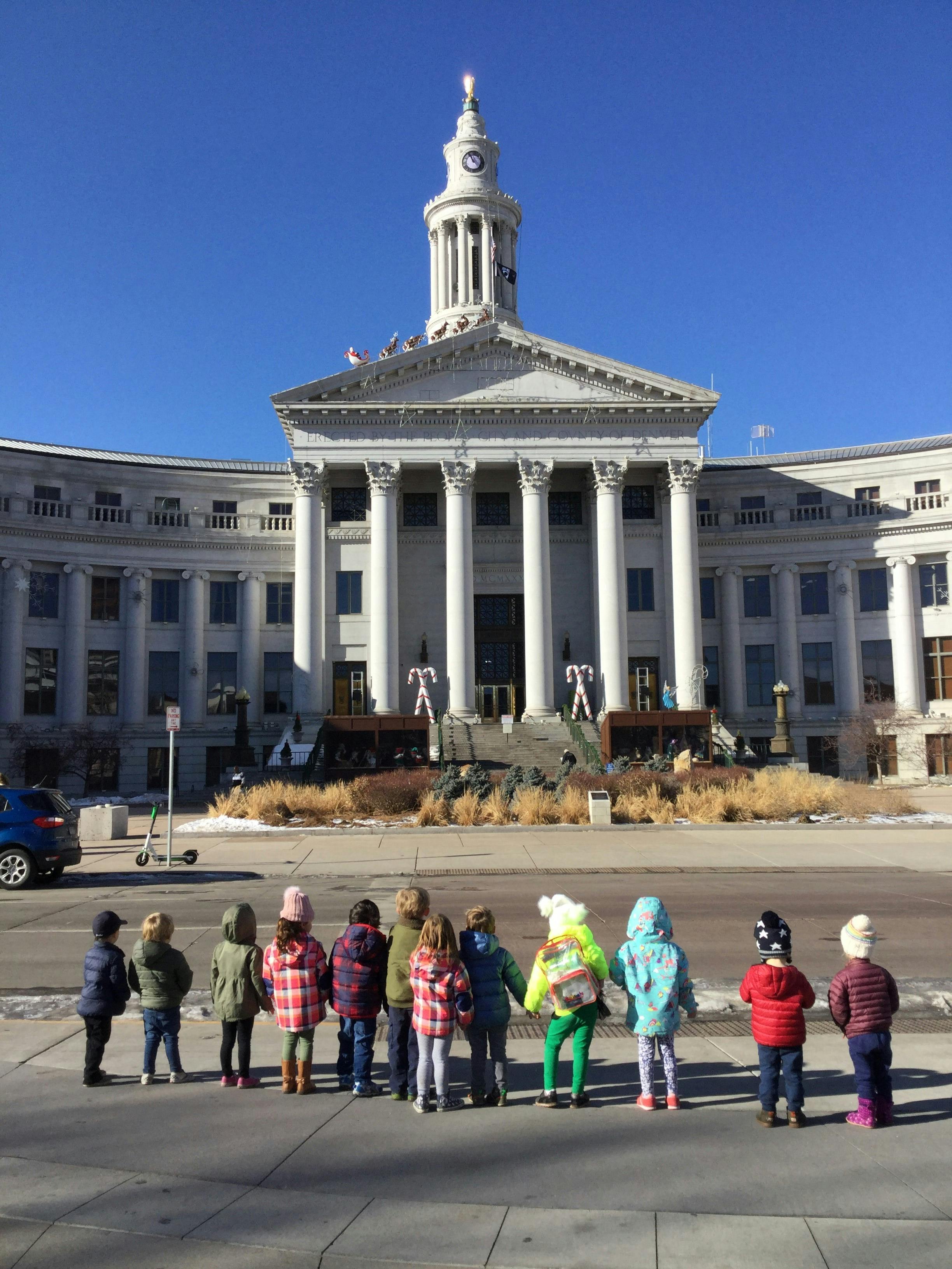Children standing on a sidewalk in front of a large building with columns and a clock tower