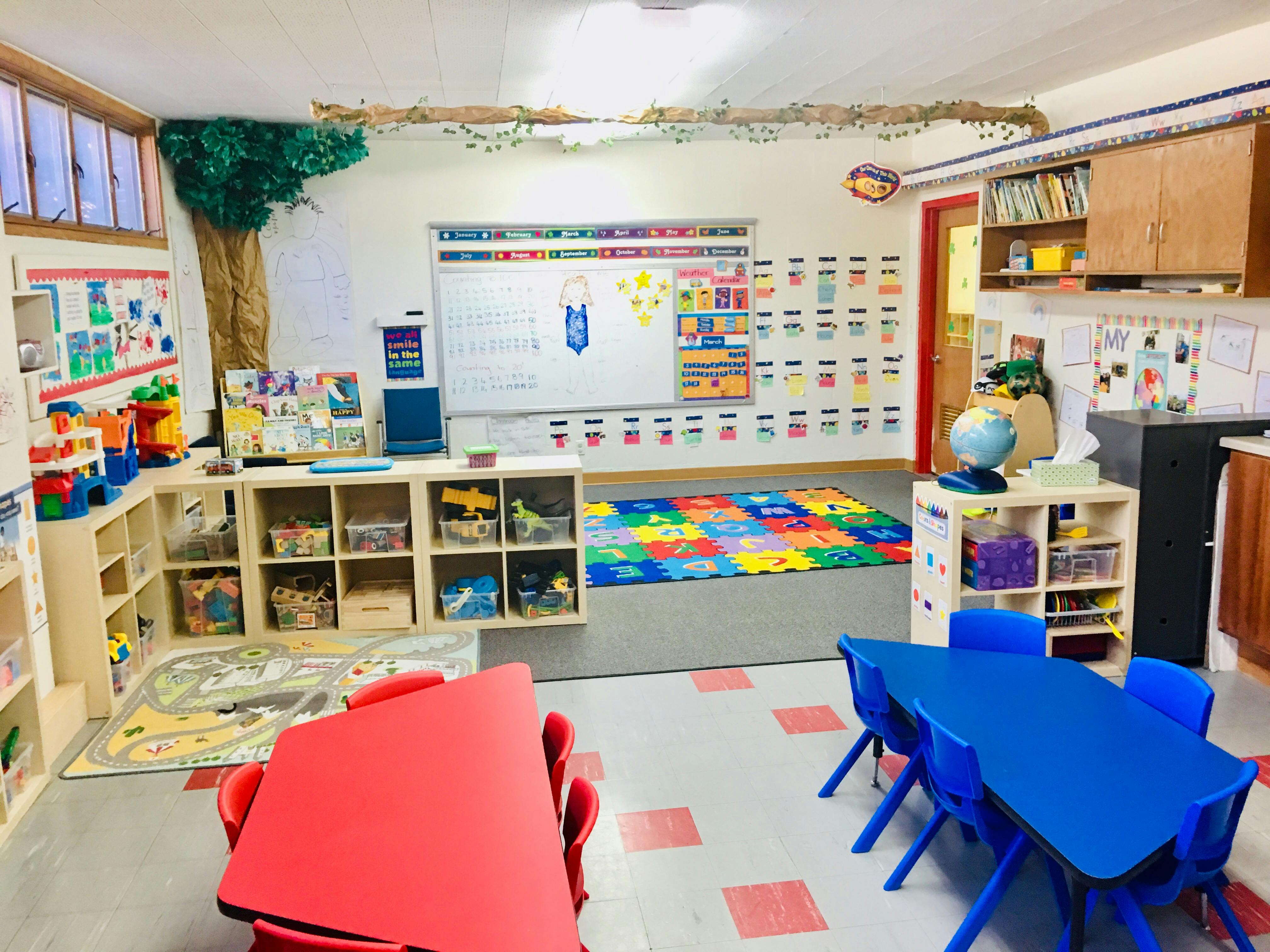 Classroom with small tables and chairs, shelves with toys, and a whiteboard