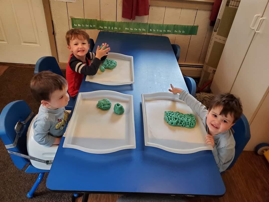 Three children playing with green dough at a blue table
