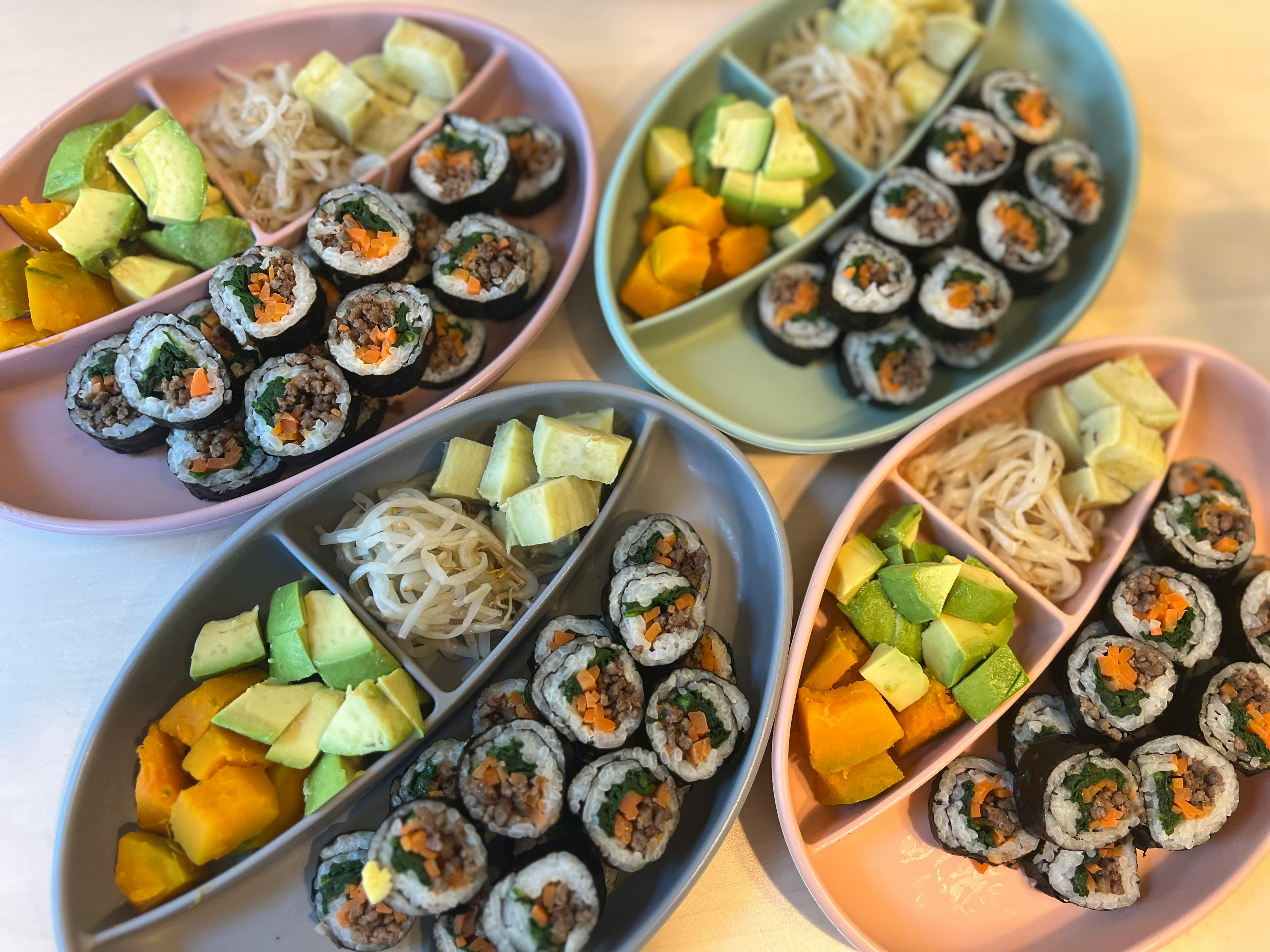 Four divided plates with sushi rolls, avocado, mango, and noodles