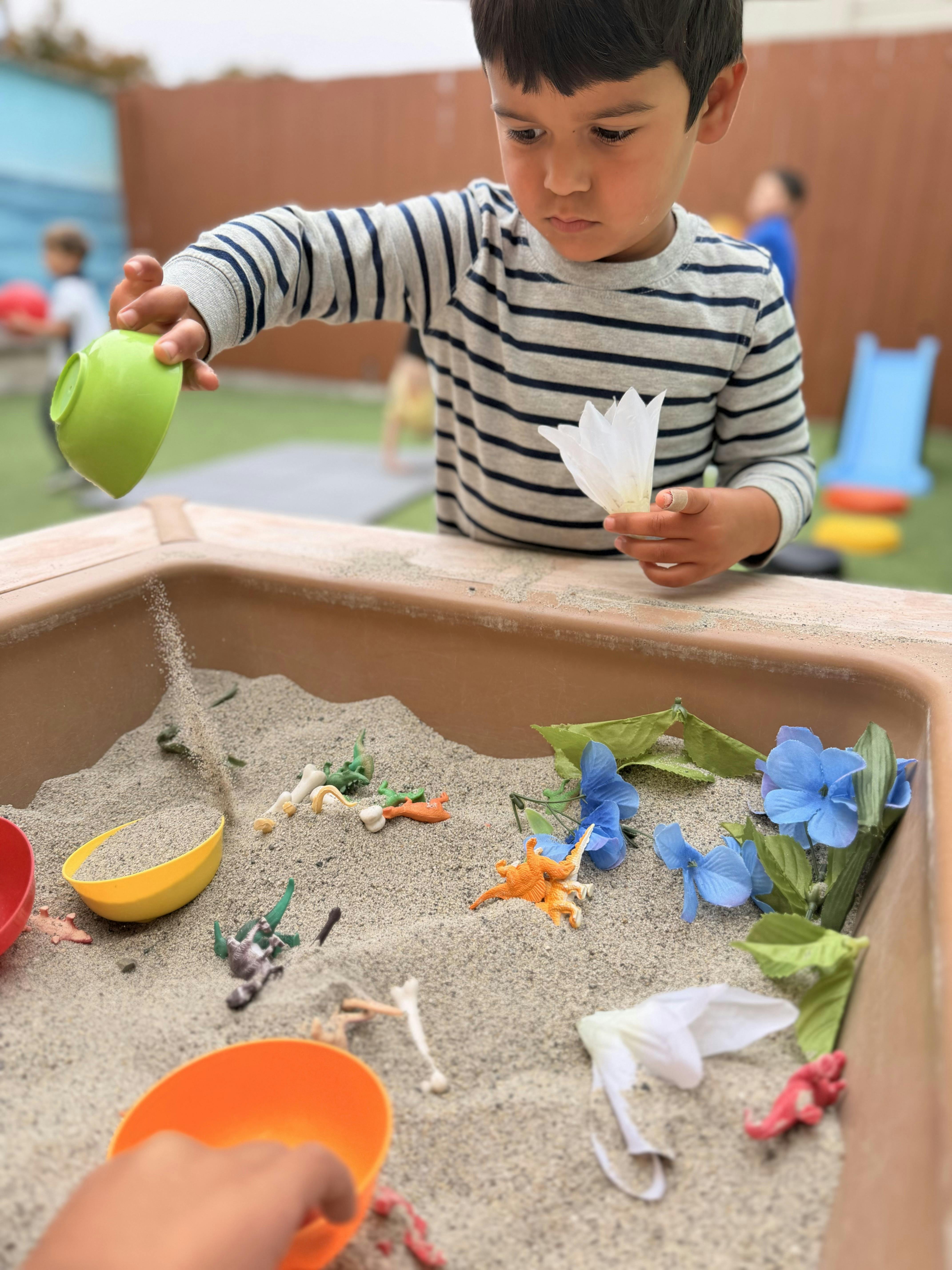 Child playing with toy dinosaurs and plastic flowers in a sandbox
