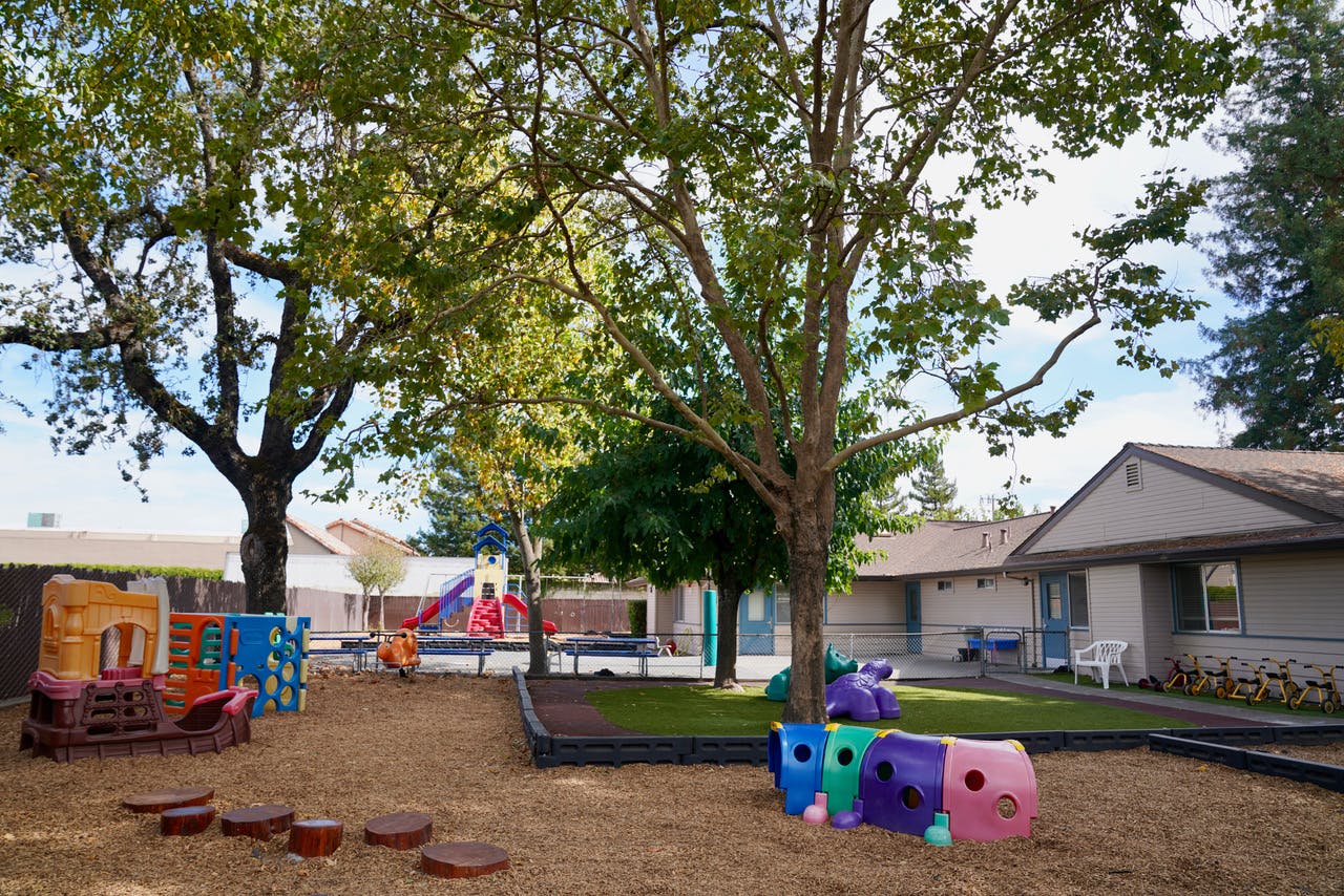 Outdoor playground with trees, play structures, and buildings in the background