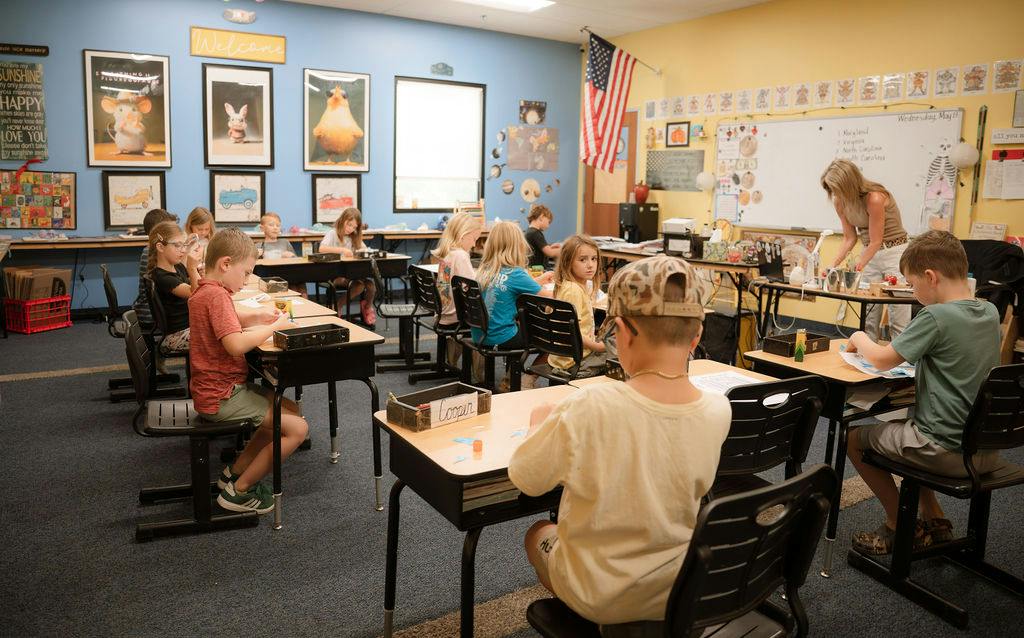 Children sitting at desks in a classroom with a teacher standing by a whiteboard