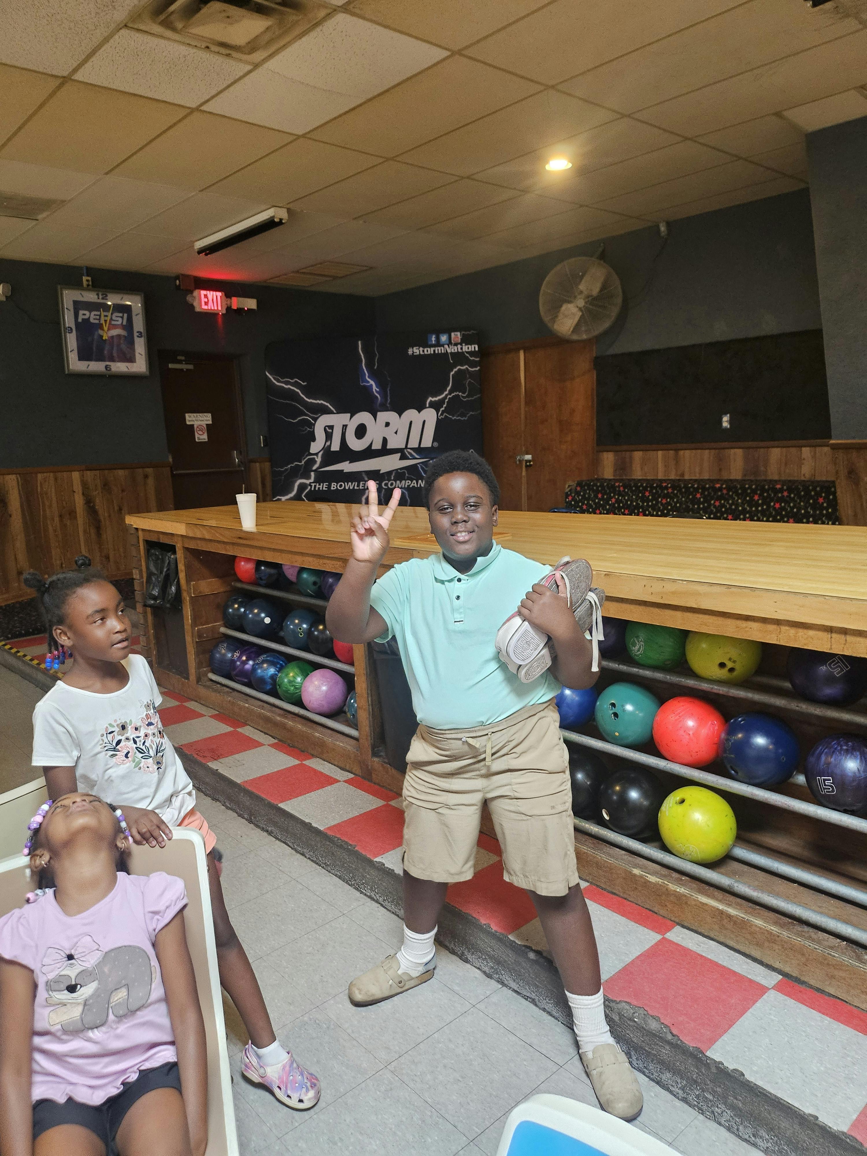 A person holding bowling shoes and making a peace sign in a bowling alley