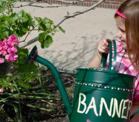 Child holding a watering can with "BANNER" written on it near pink flowers