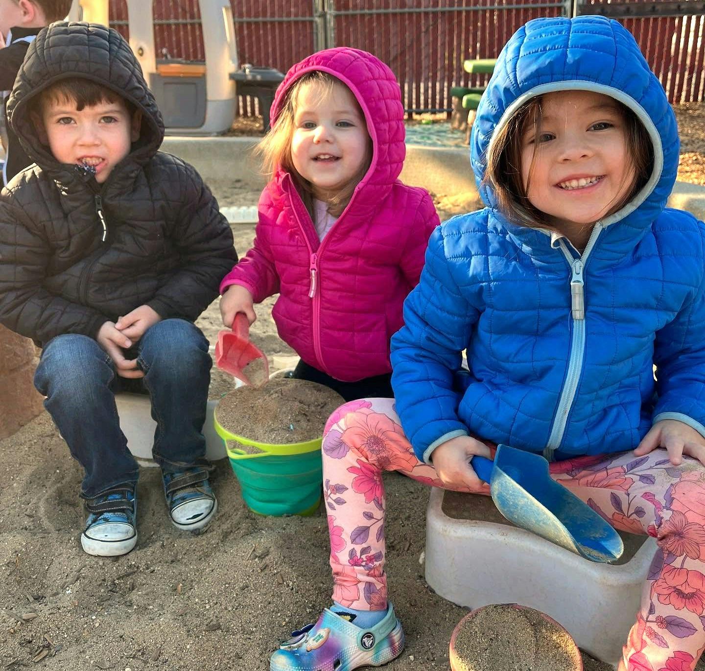 Three children in winter coats playing in a sandbox