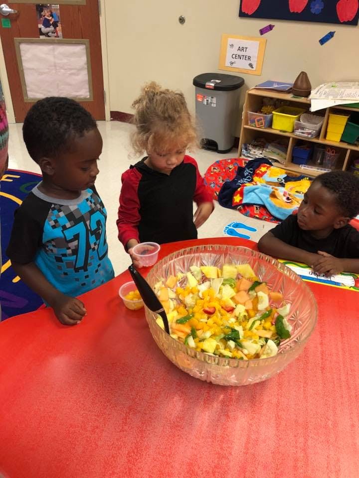 Three children looking at a large bowl of fruit salad on a red table