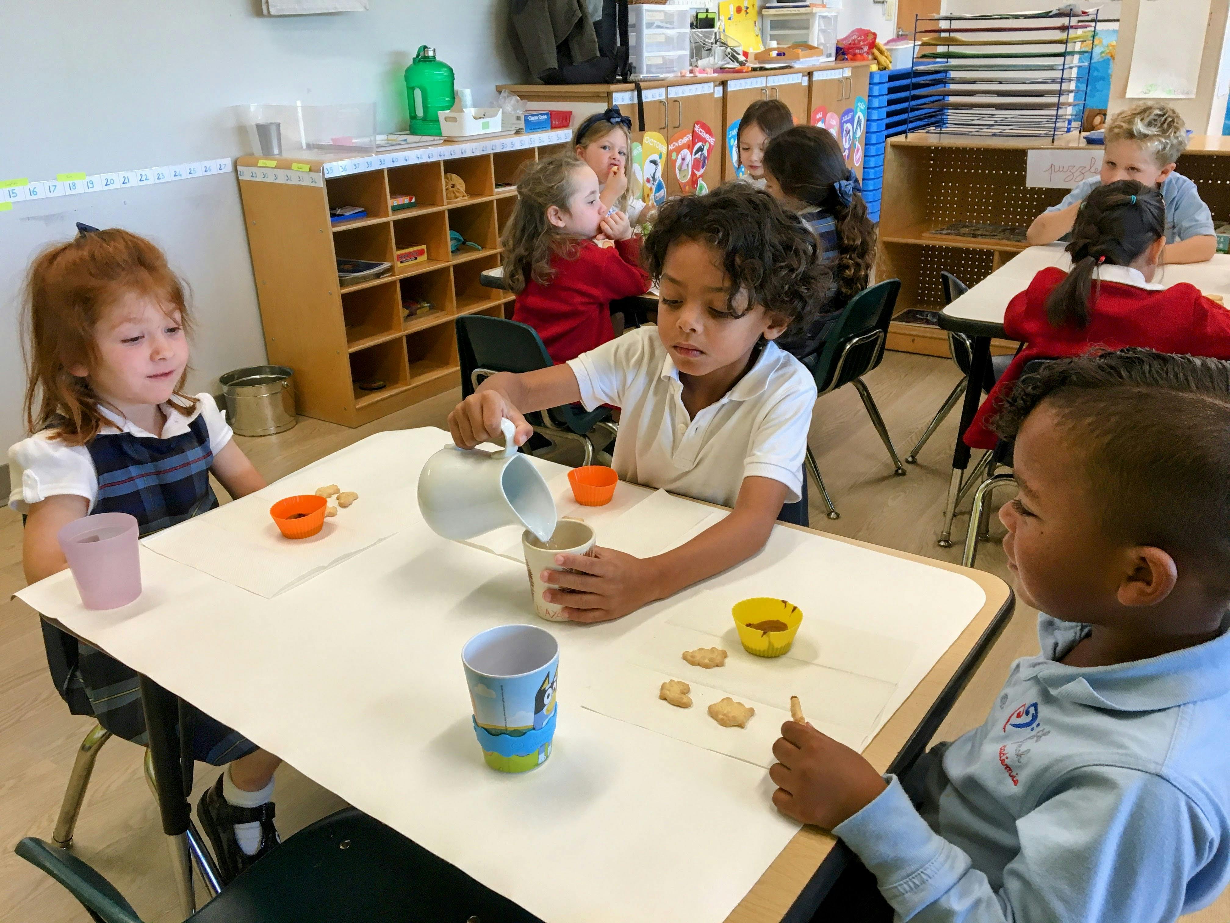 Child pouring from a pitcher into a cup at a table with other children