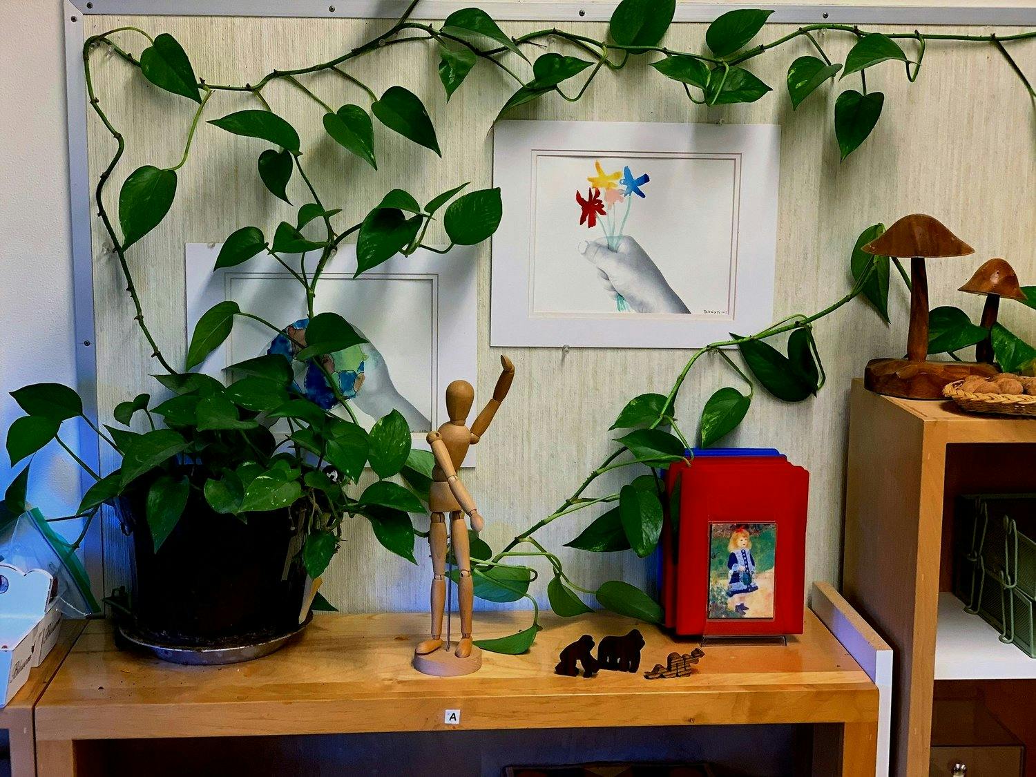 Potted plant and wooden mannequin on a shelf with artwork and vines on the wall