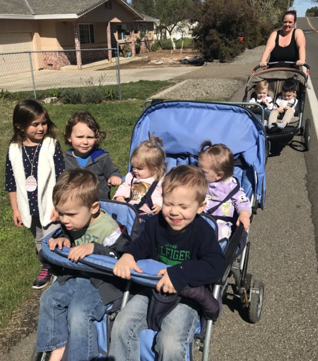 Person pushing two strollers with multiple children on a sidewalk