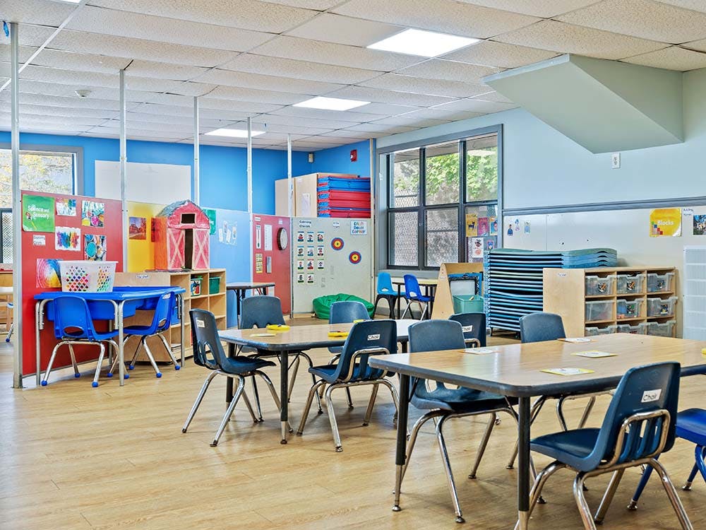 Empty classroom with small tables and chairs, colorful dividers, and windows
