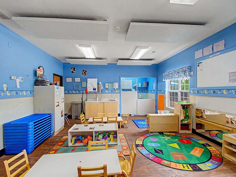 Empty children's classroom with small tables and chairs, a play rug, and stacked cots