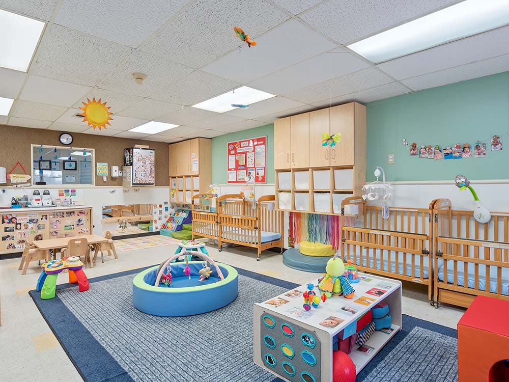 A colorful daycare room with cribs, play mats, and small tables with chairs
