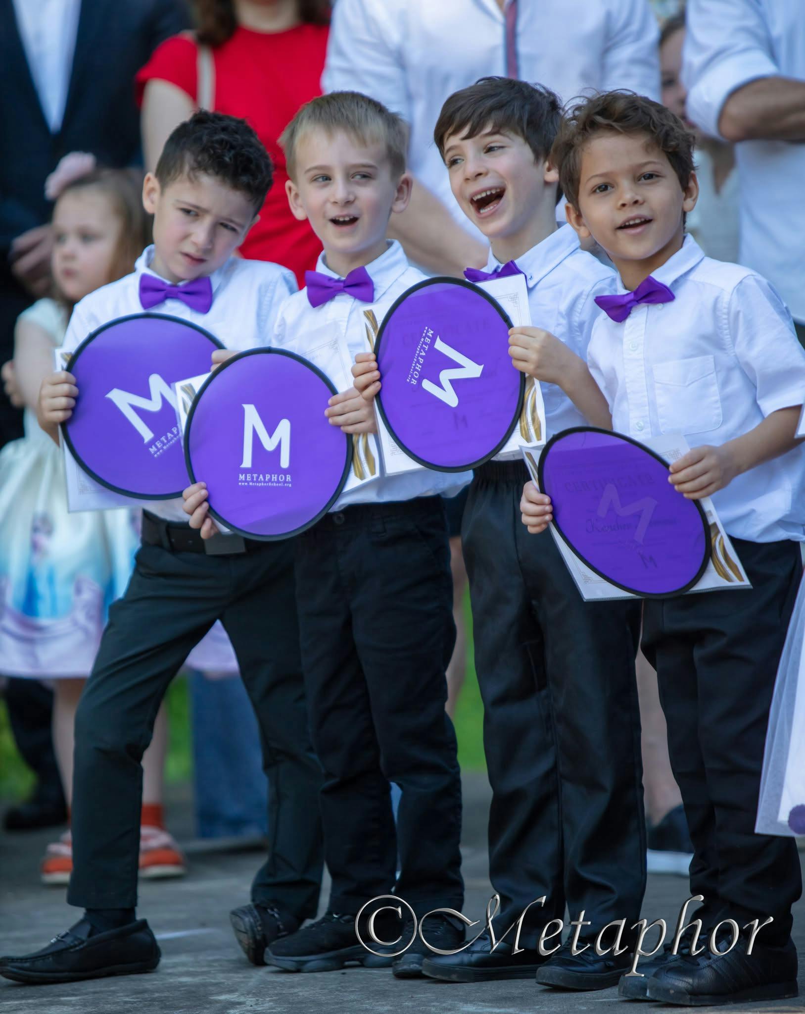 Four children in white shirts and purple bow ties holding purple circles with an M
