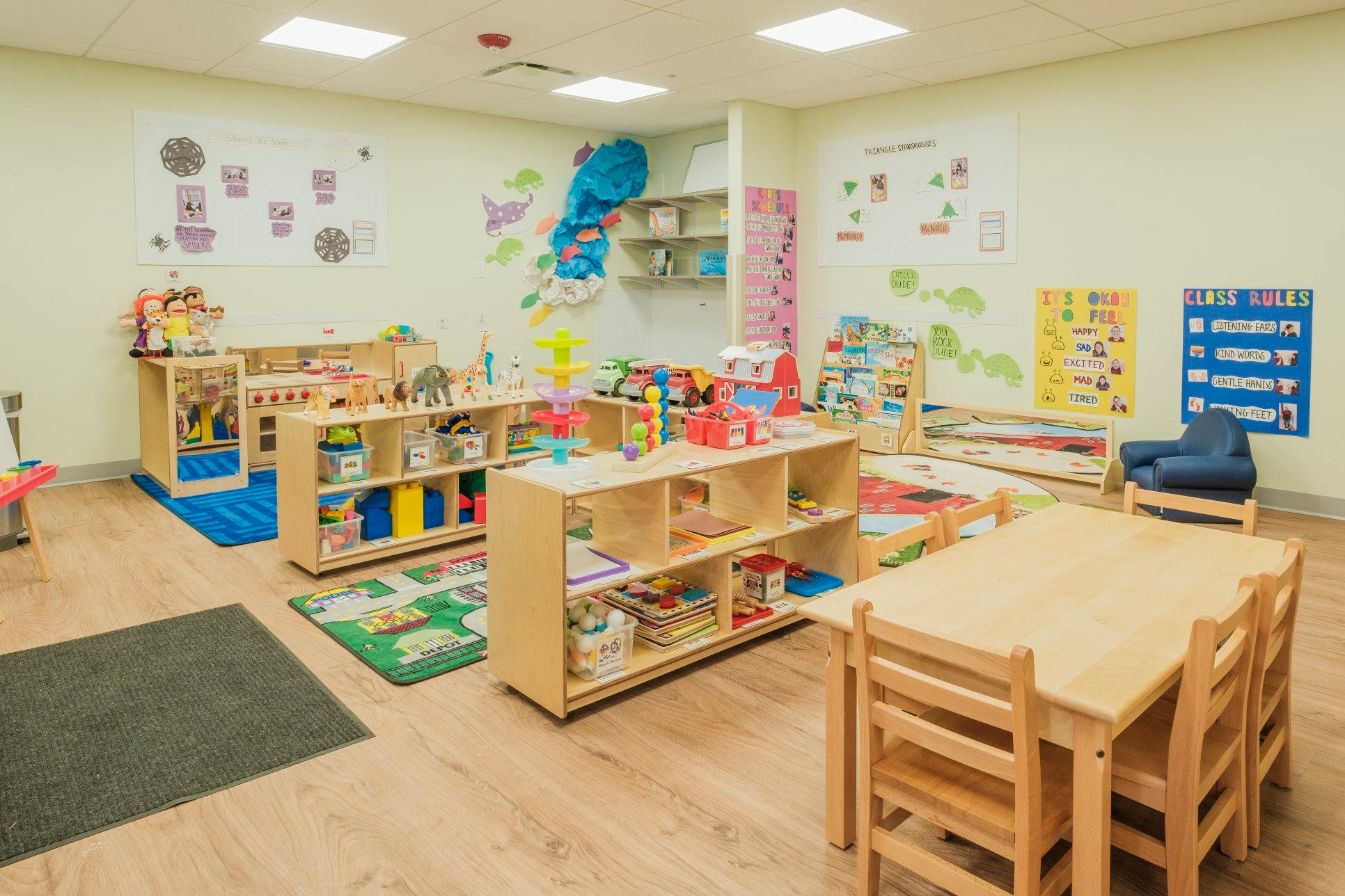 An empty classroom with wooden tables and chairs, shelves full of toys and books