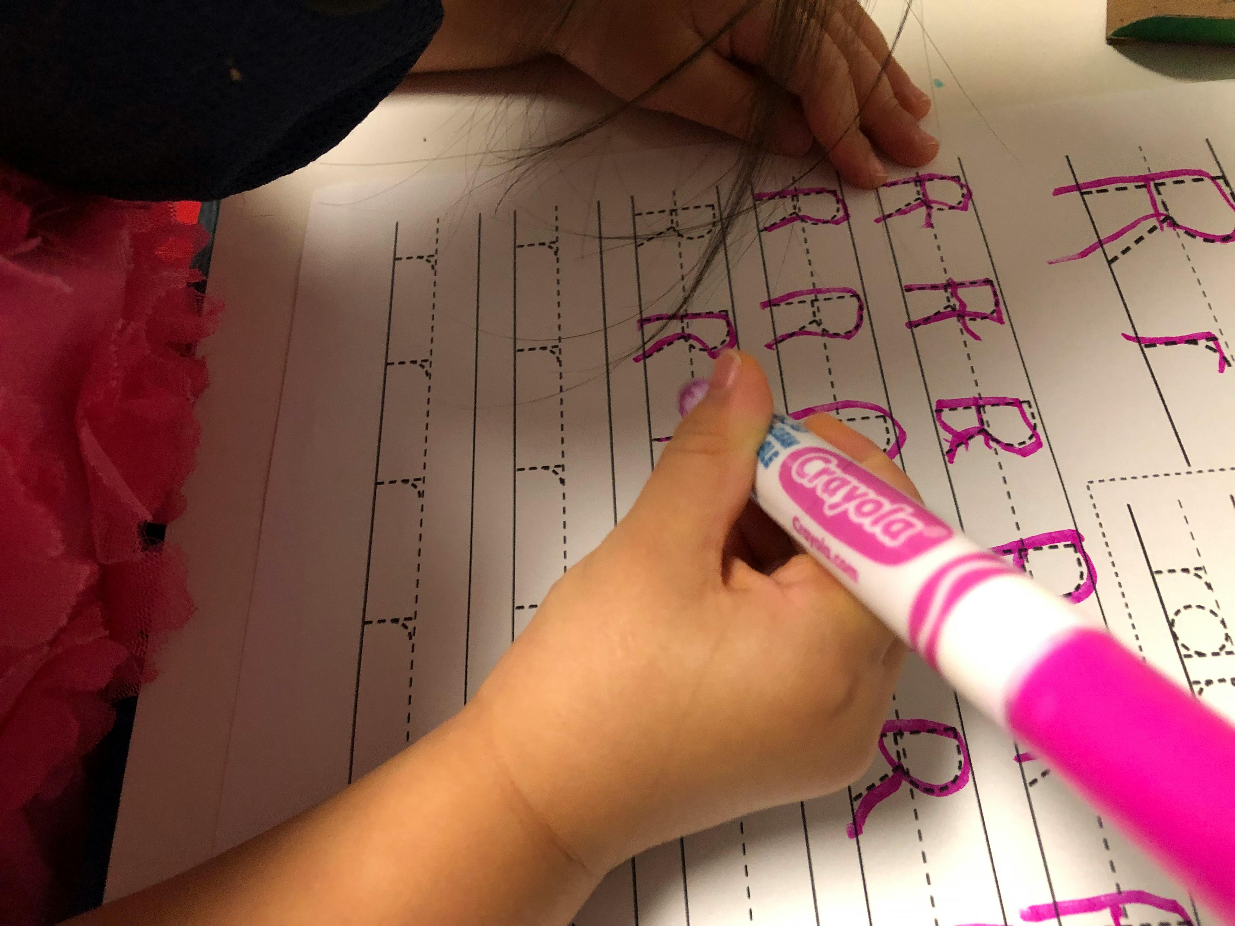 Child's hand holding a pink marker, tracing letters on paper
