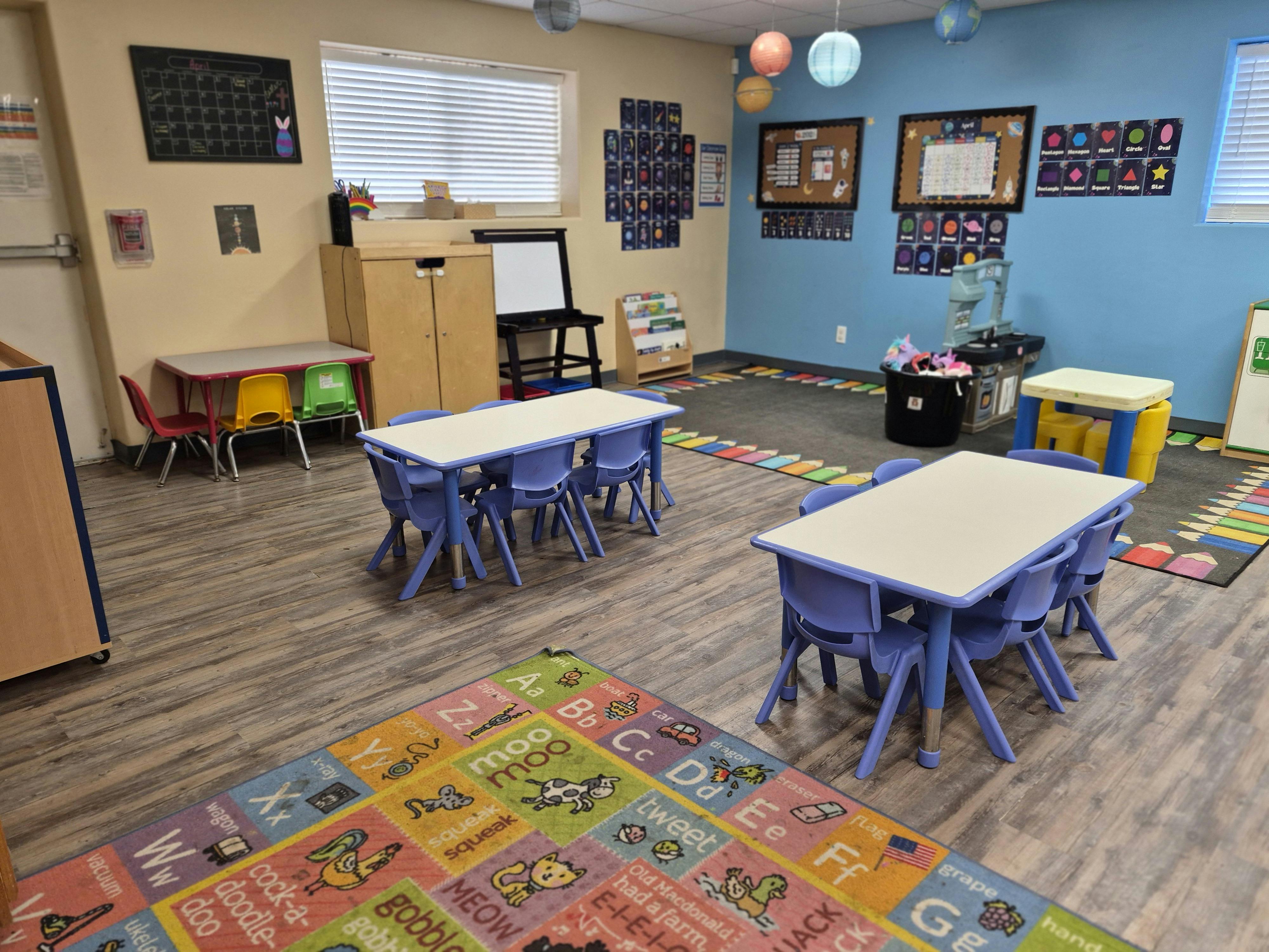 Classroom with blue tables, colorful chairs, educational posters on walls, and an alphabet rug