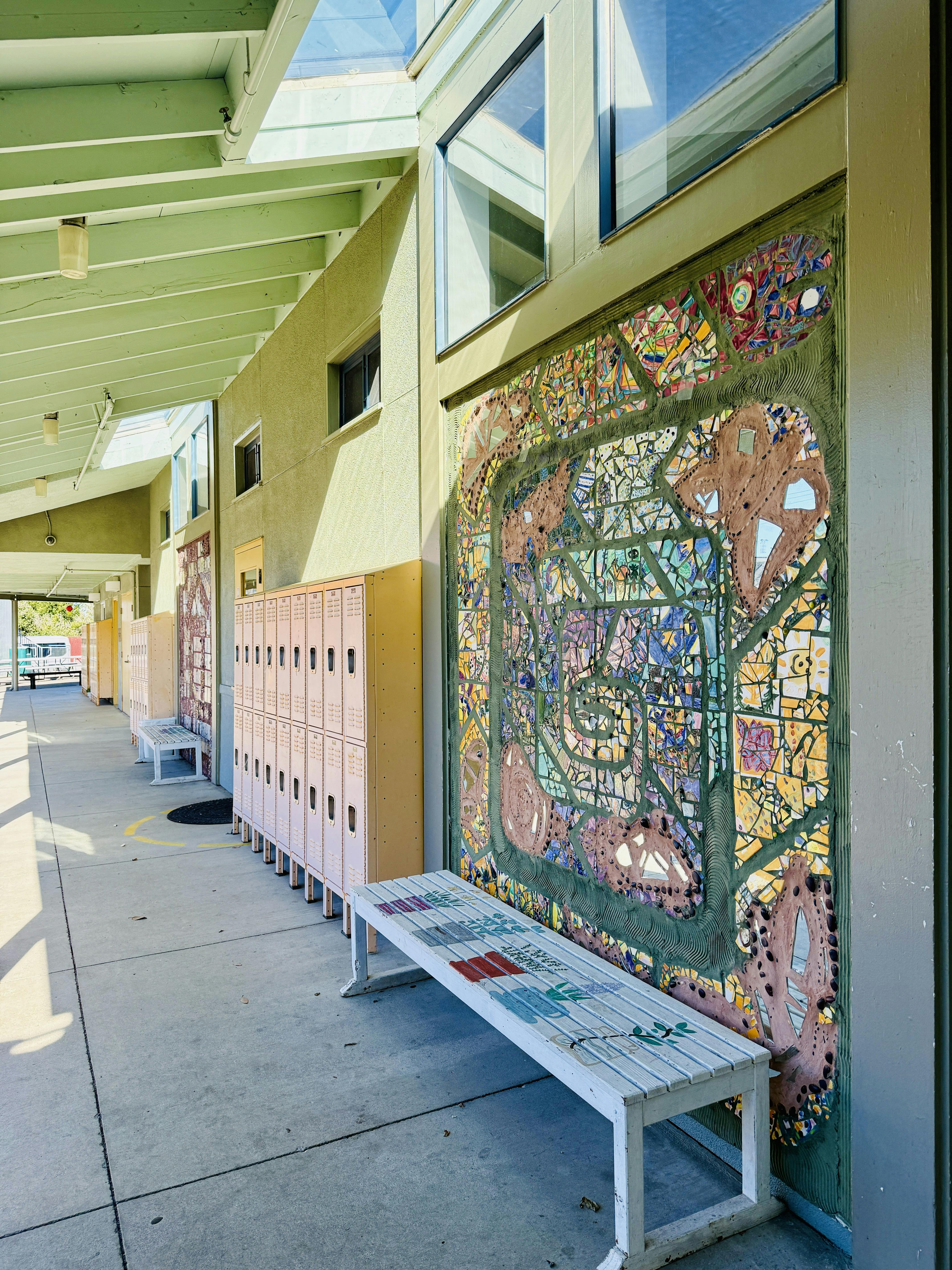 Colorful mosaic wall art and benches along an outdoor school hallway with lockers