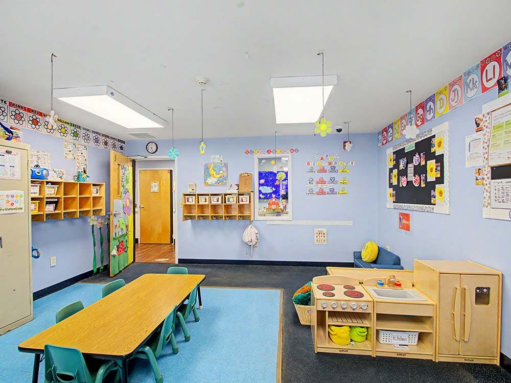 An empty children's classroom with a long table, chairs, and play kitchen set