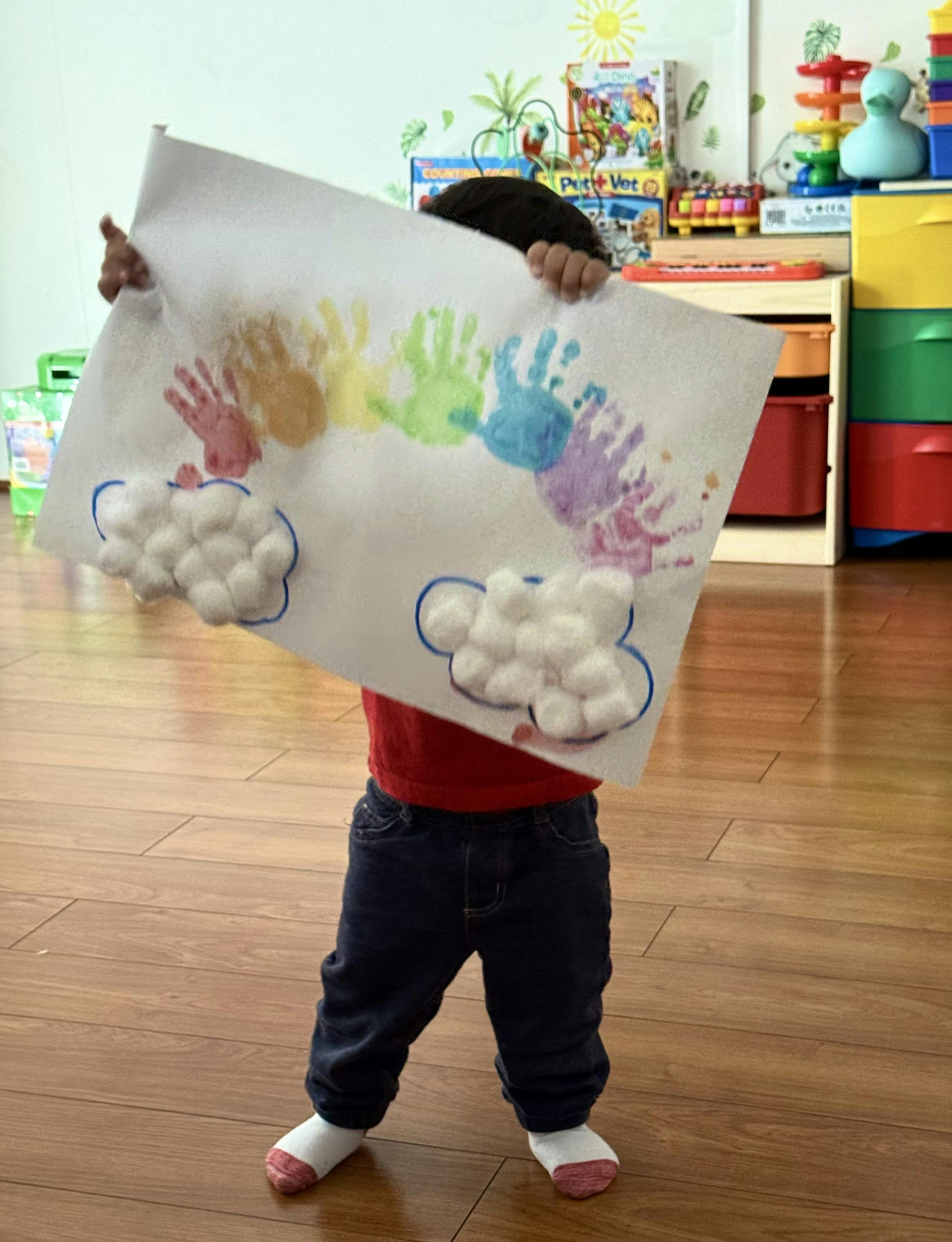 Child holding up paper with rainbow handprints and cotton ball clouds