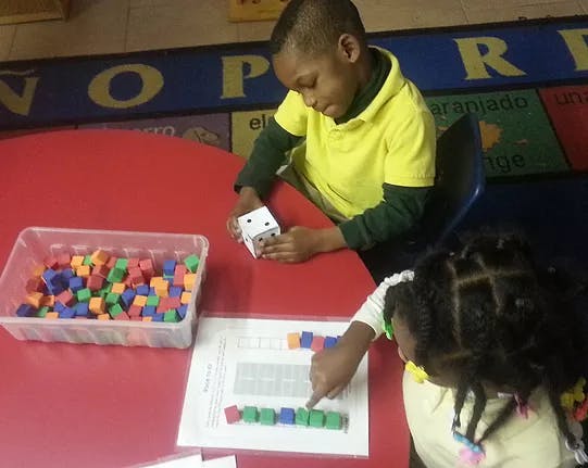 Two children playing with colorful blocks and a die at a red table