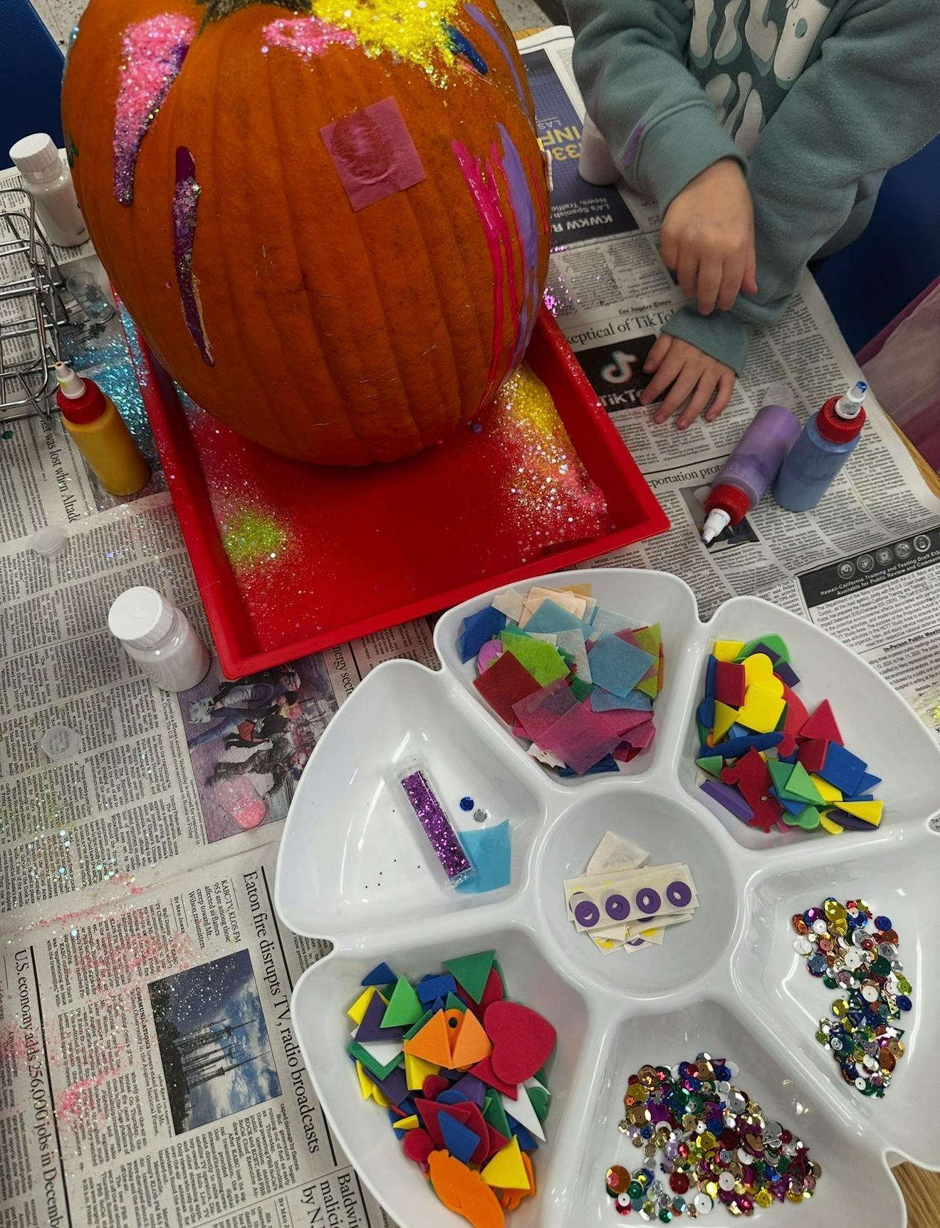 Child decorating a pumpkin with glitter and glue, surrounded by craft supplies