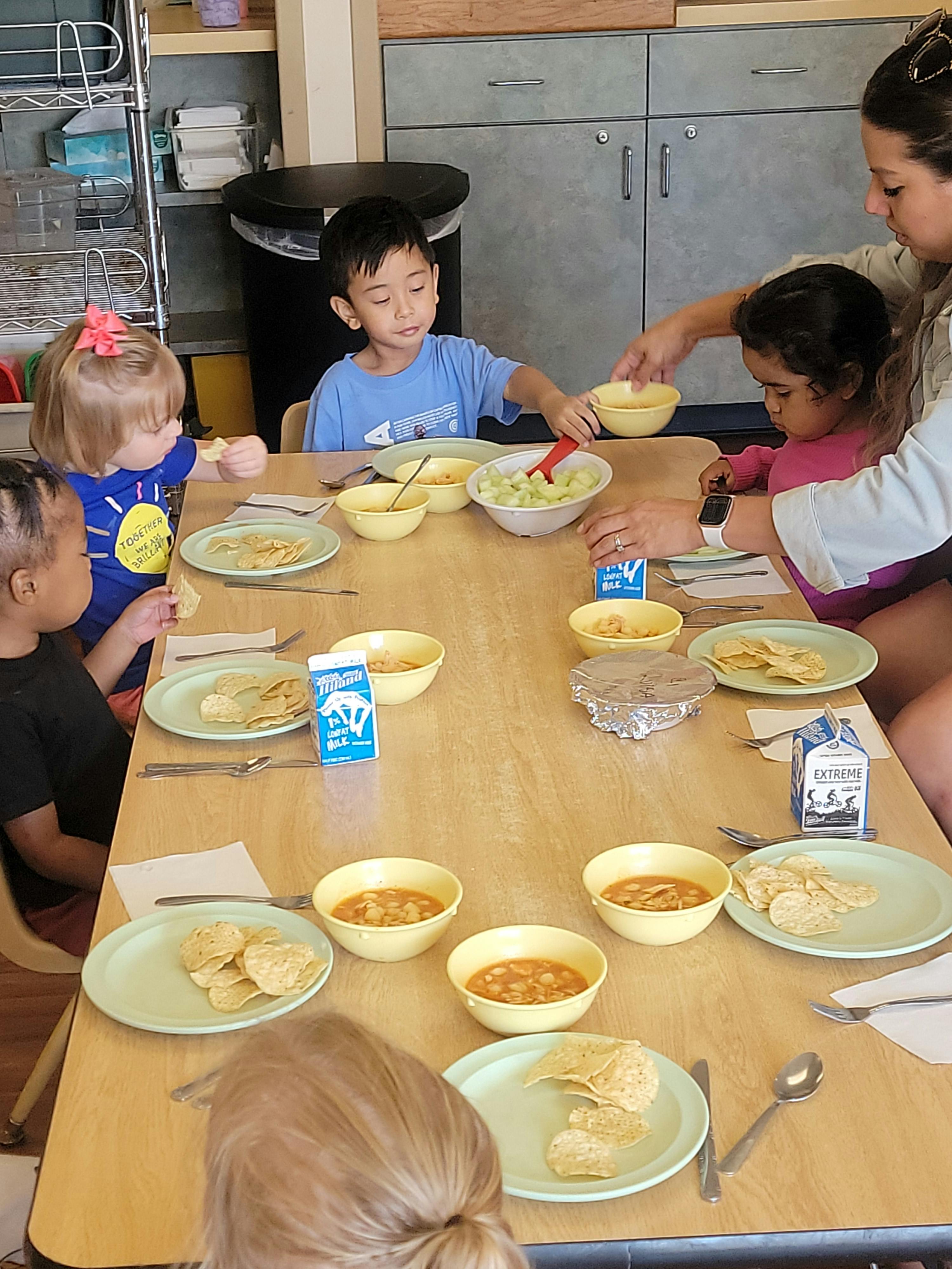 Adult serving food to children sitting at a table with plates of chips and soup
