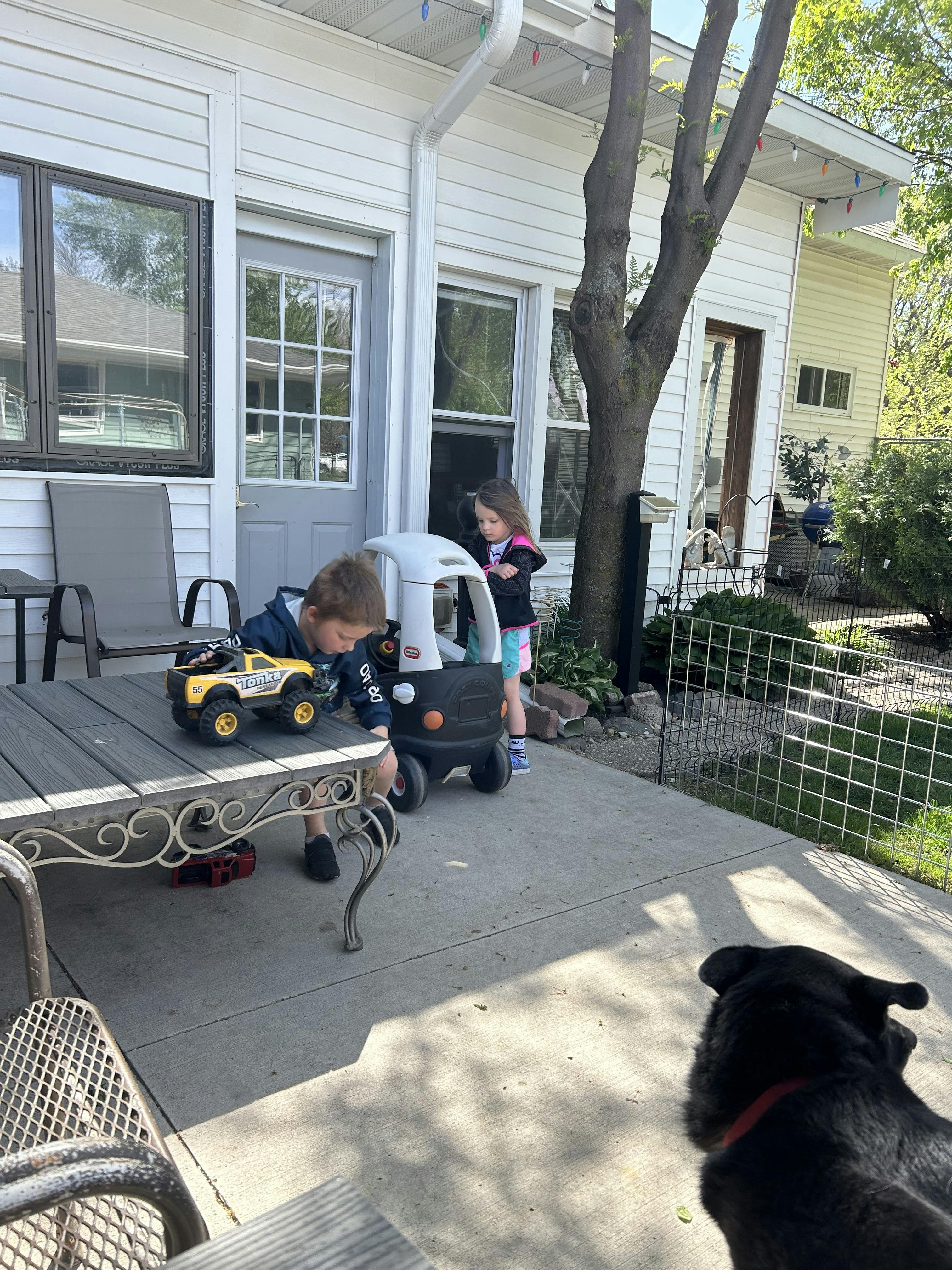child playing with a yellow toy truck on a patio table, another child next to a toy car
