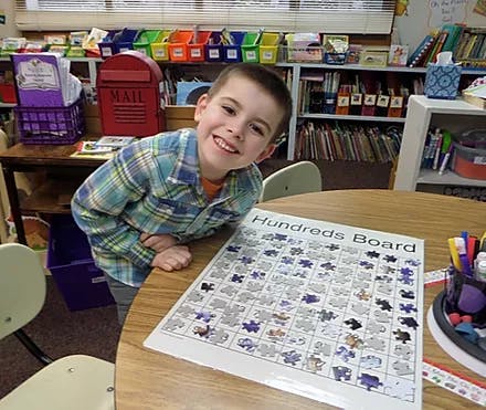 Child leaning on a table with a "Hundreds Board" in a classroom