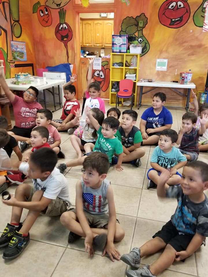 Children sitting on the floor in a classroom, some raising their hands