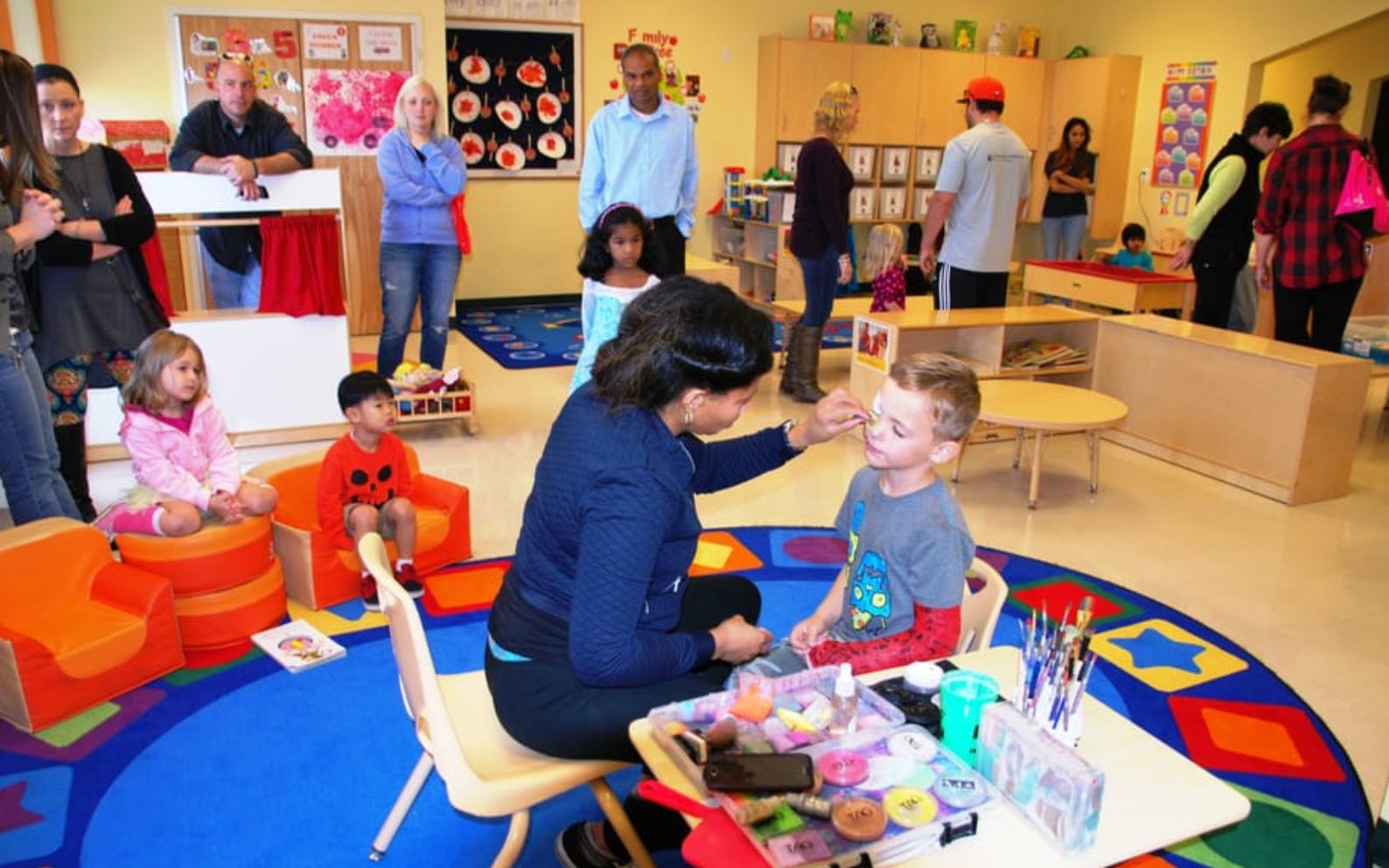 Adult applying makeup to a child in a classroom with other adults and children