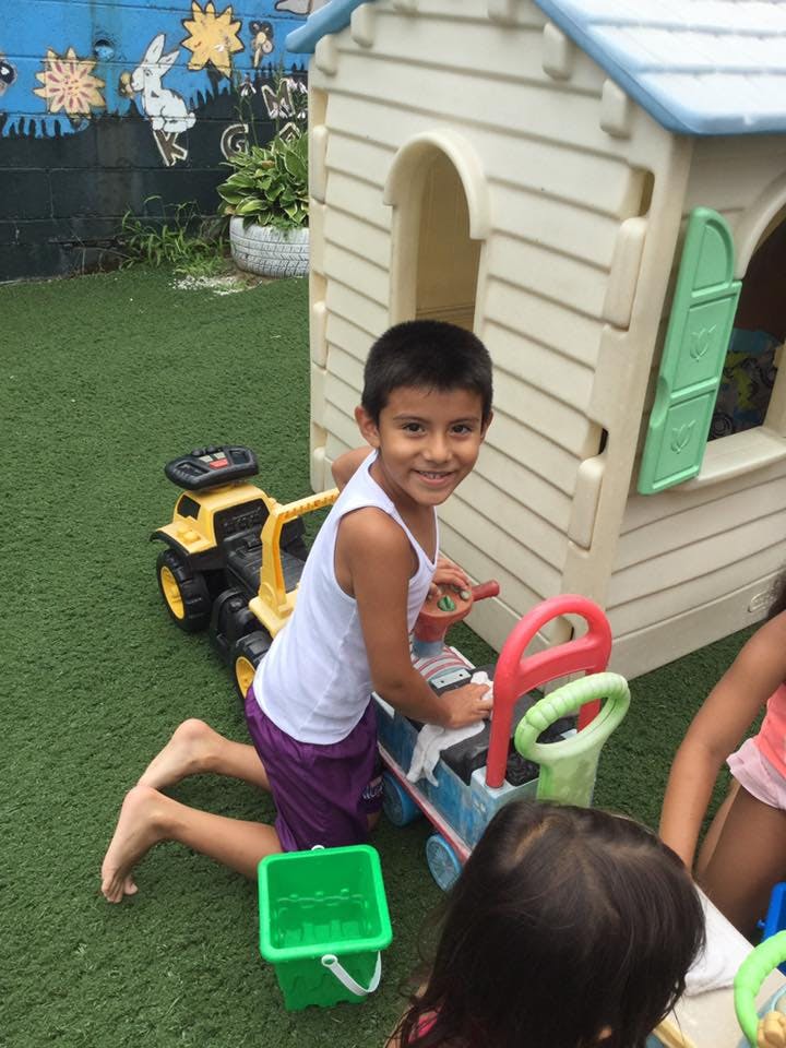 Child kneeling on artificial grass next to a toy car and playhouse