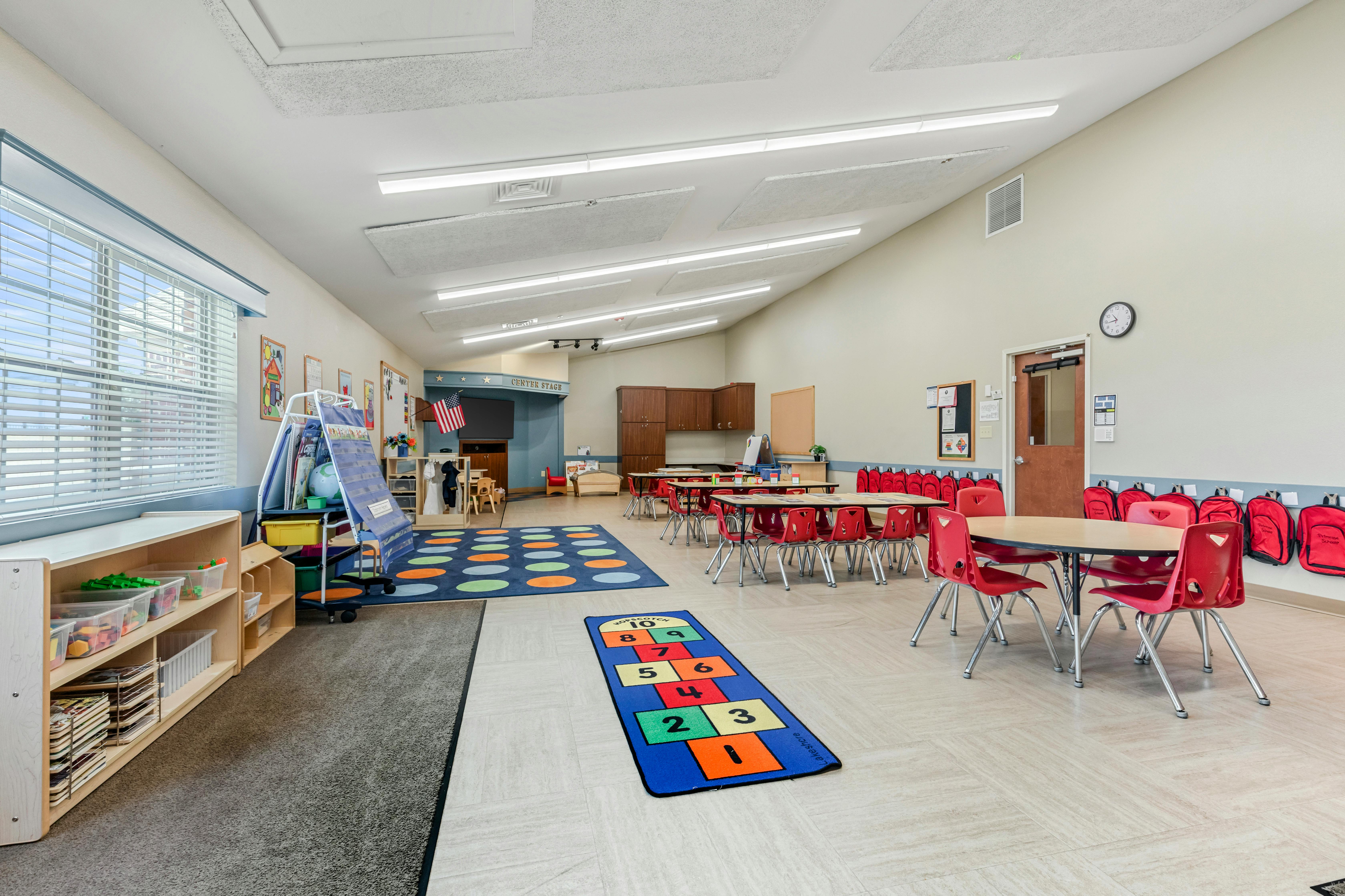 An empty classroom with small red chairs, tables, and play mats