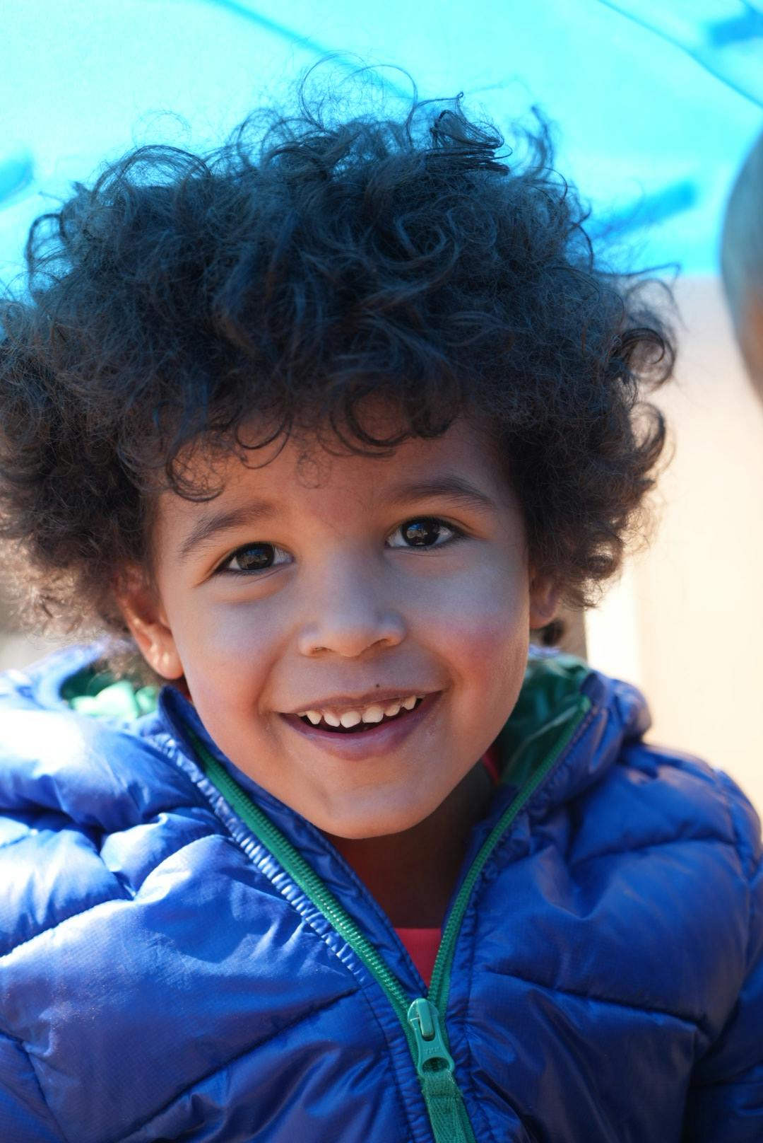 Child in a blue winter coat with dark curly hair smiling directly at the camera