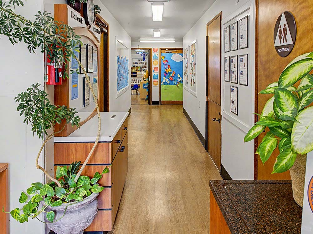 Hallway with wood floors, plants, a restroom sign, and a children's classroom