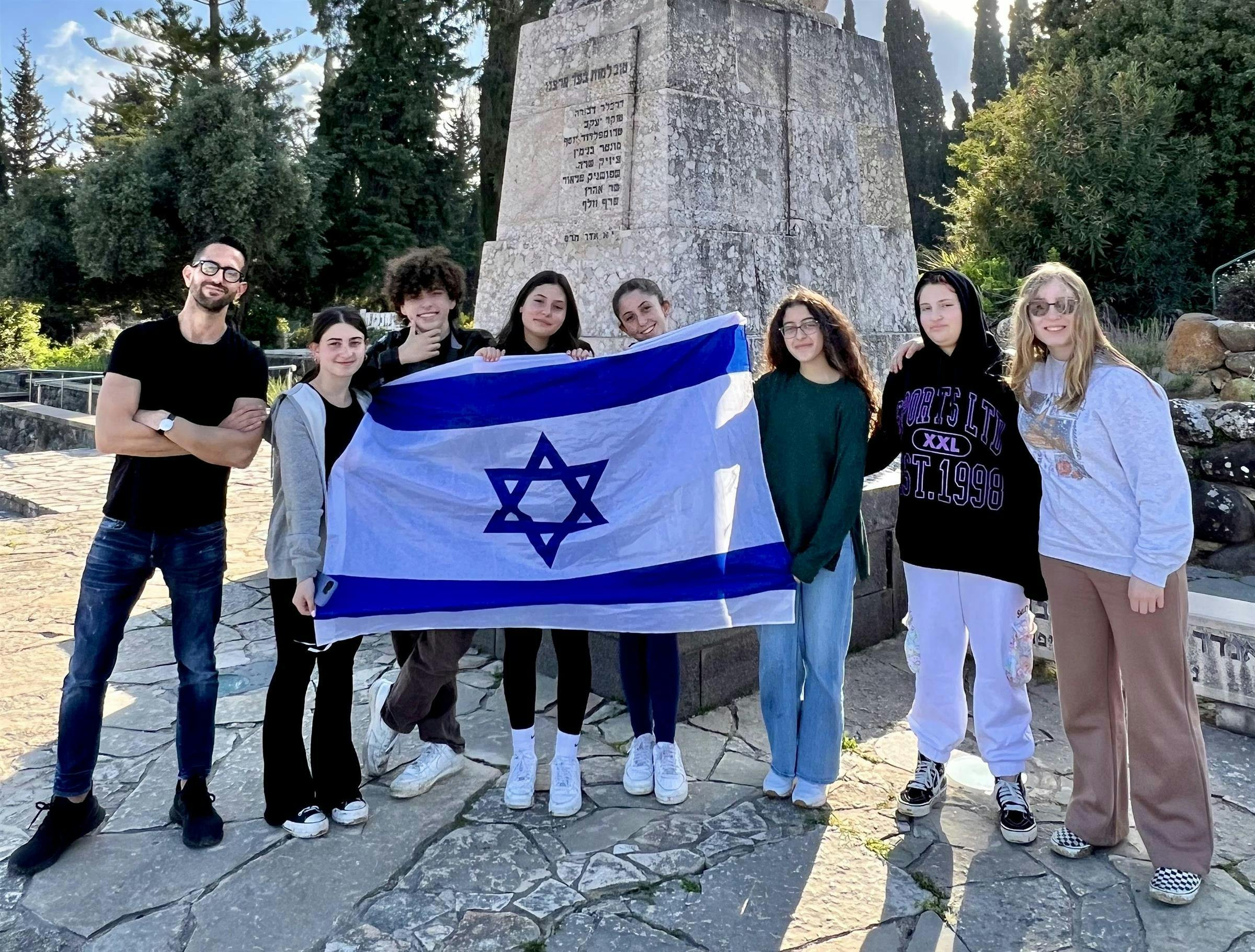 Group of adults and teens holding an Israeli flag in front of a stone monument