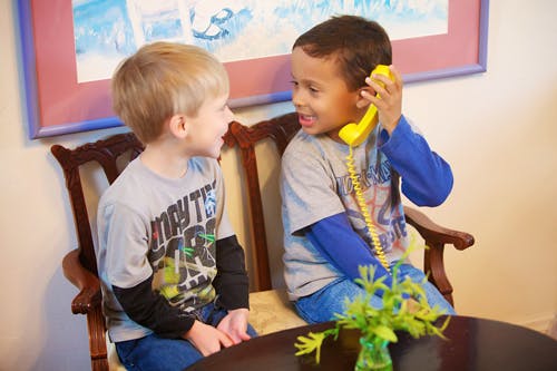 Two children sitting in chairs, one holding a yellow phone to an ear