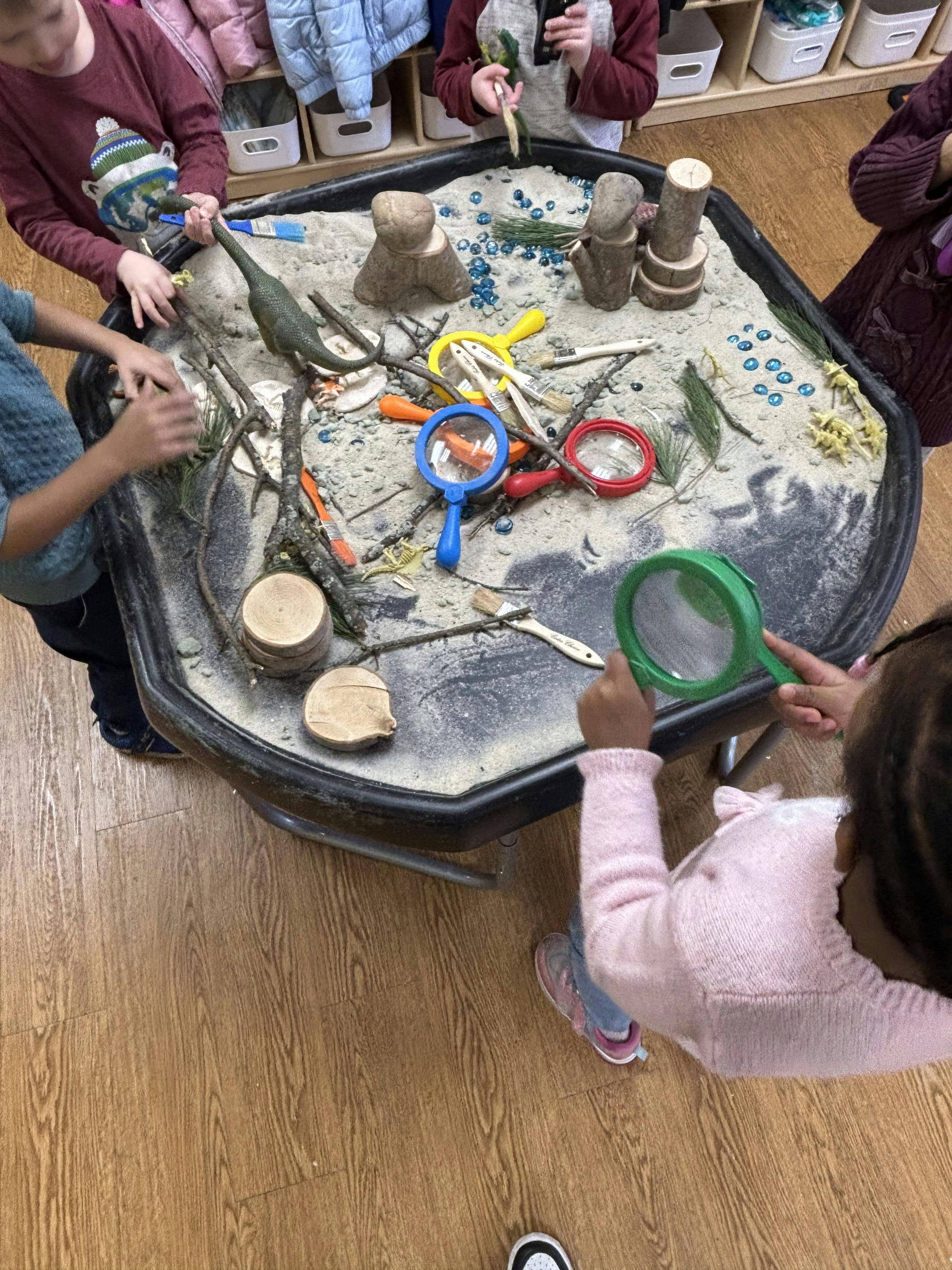 Children playing around a sensory bin filled with sand, magnifying glasses, and dinosaur toys