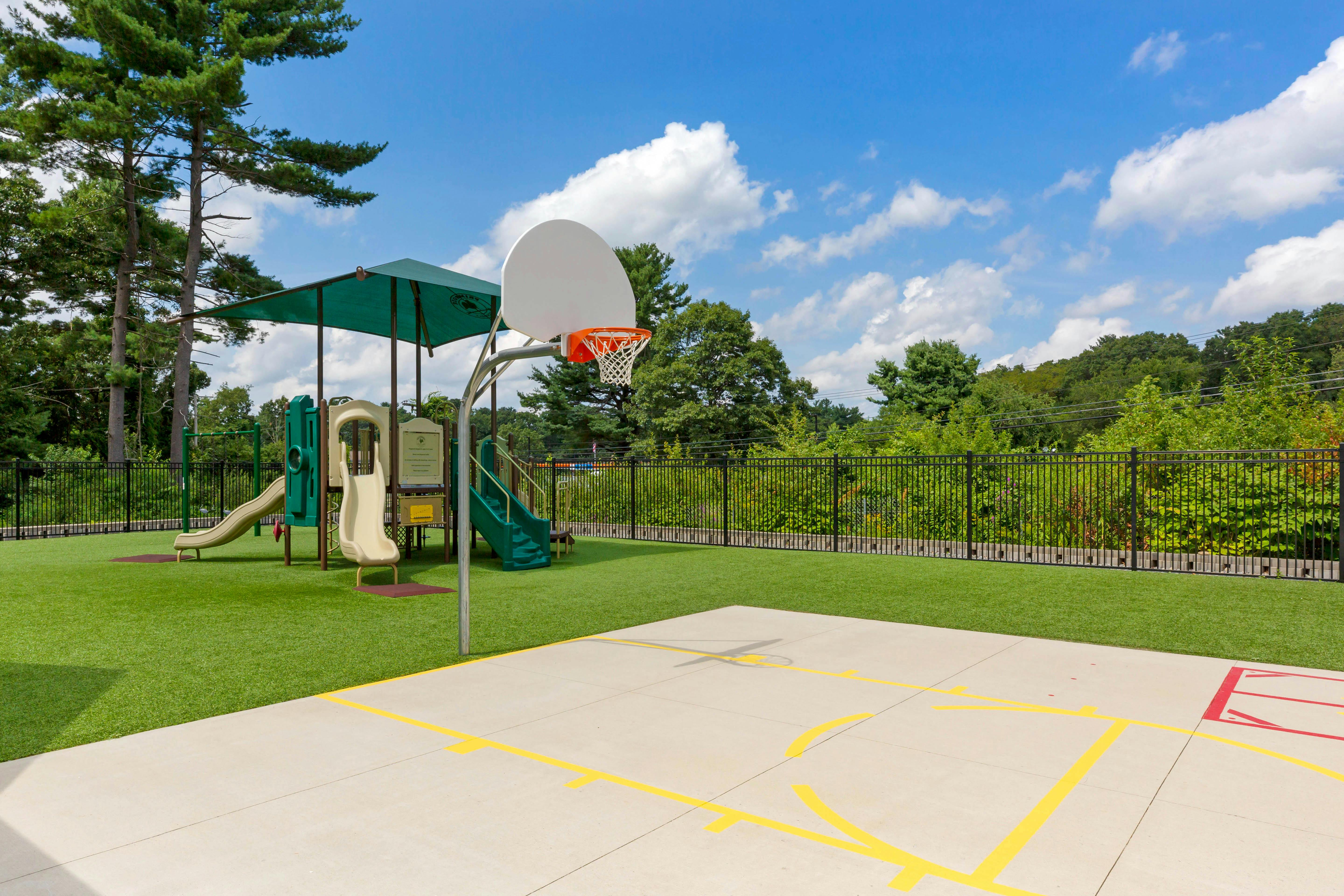 Basketball court and playground set on a green turf surrounded by a black fence