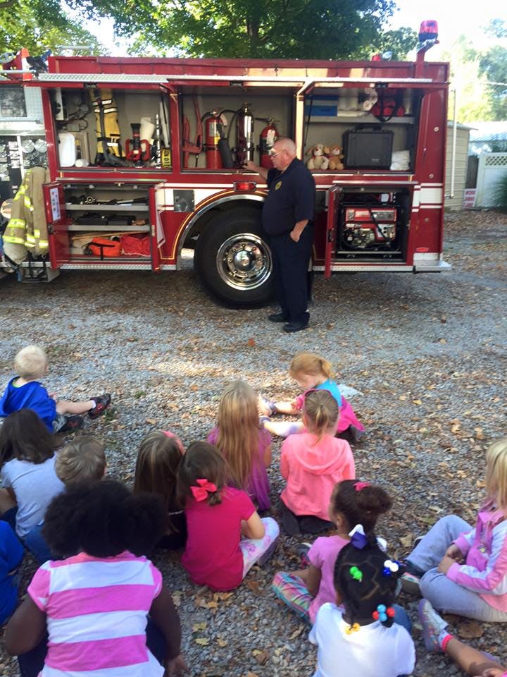 Firefighter standing by a fire truck with children sitting on the ground