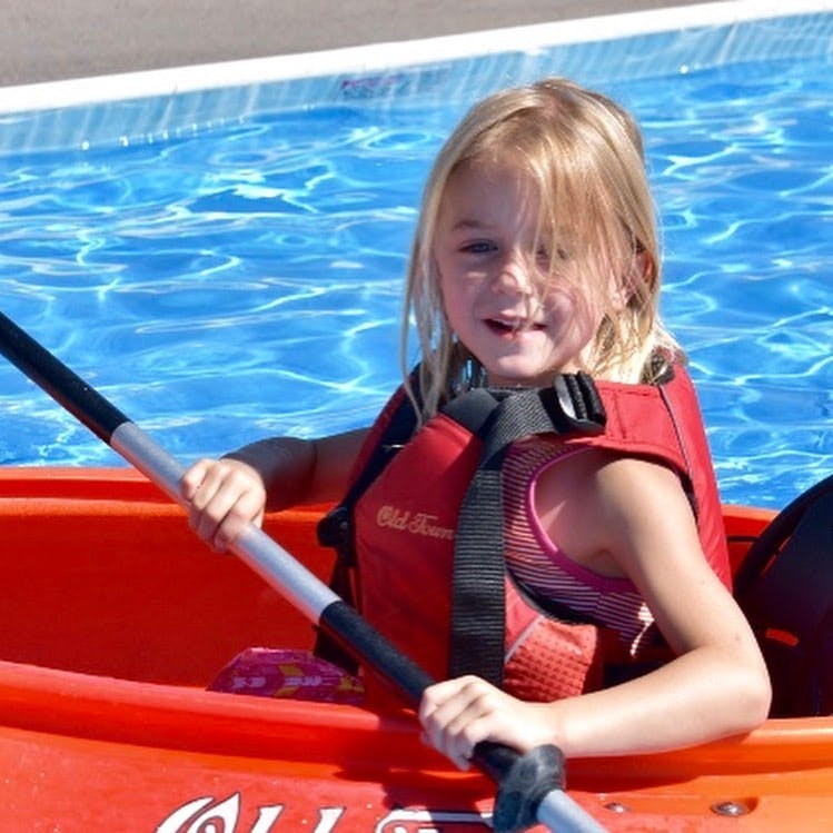 Child in a red life vest holding a paddle in a kayak in a swimming pool