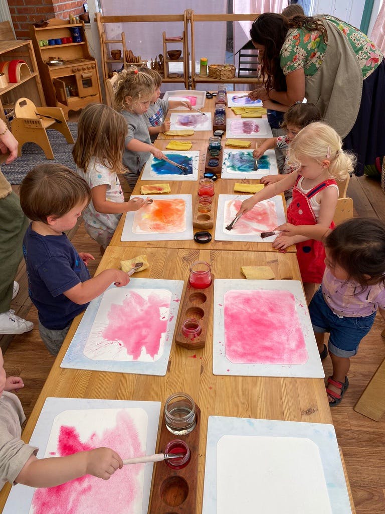Several children watercolor painting at a long wooden table with an adult present