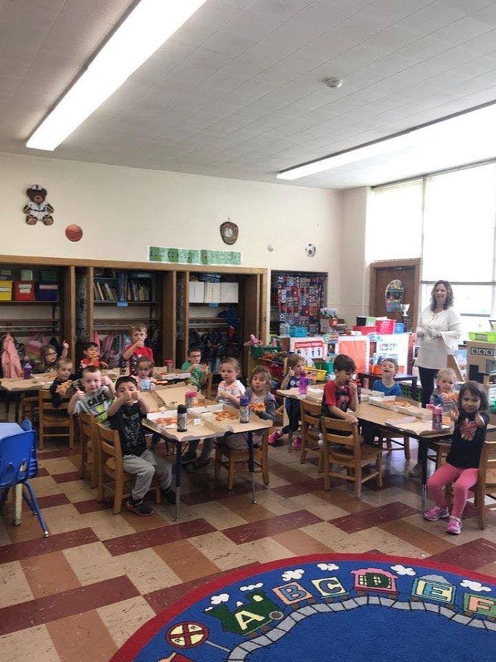 Children sitting at tables in a classroom with an adult standing