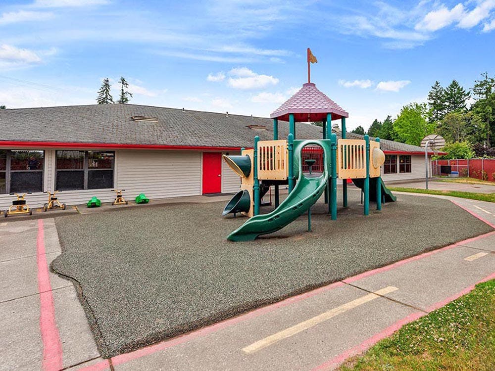 Playground with green and tan play structure, slides, and red roof building