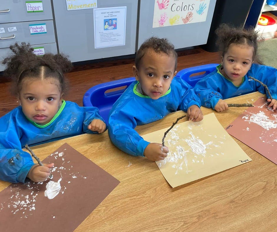 Three children in blue smocks painting with sticks in a classroom