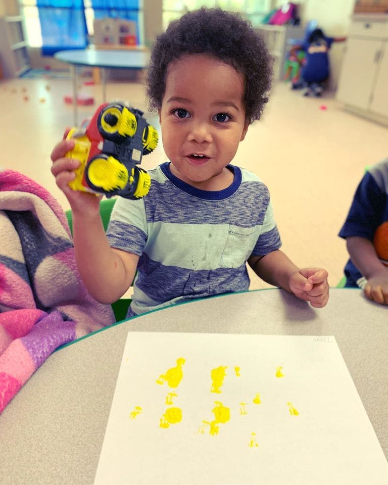 Child holding a toy and looking at the camera, with yellow paint on paper