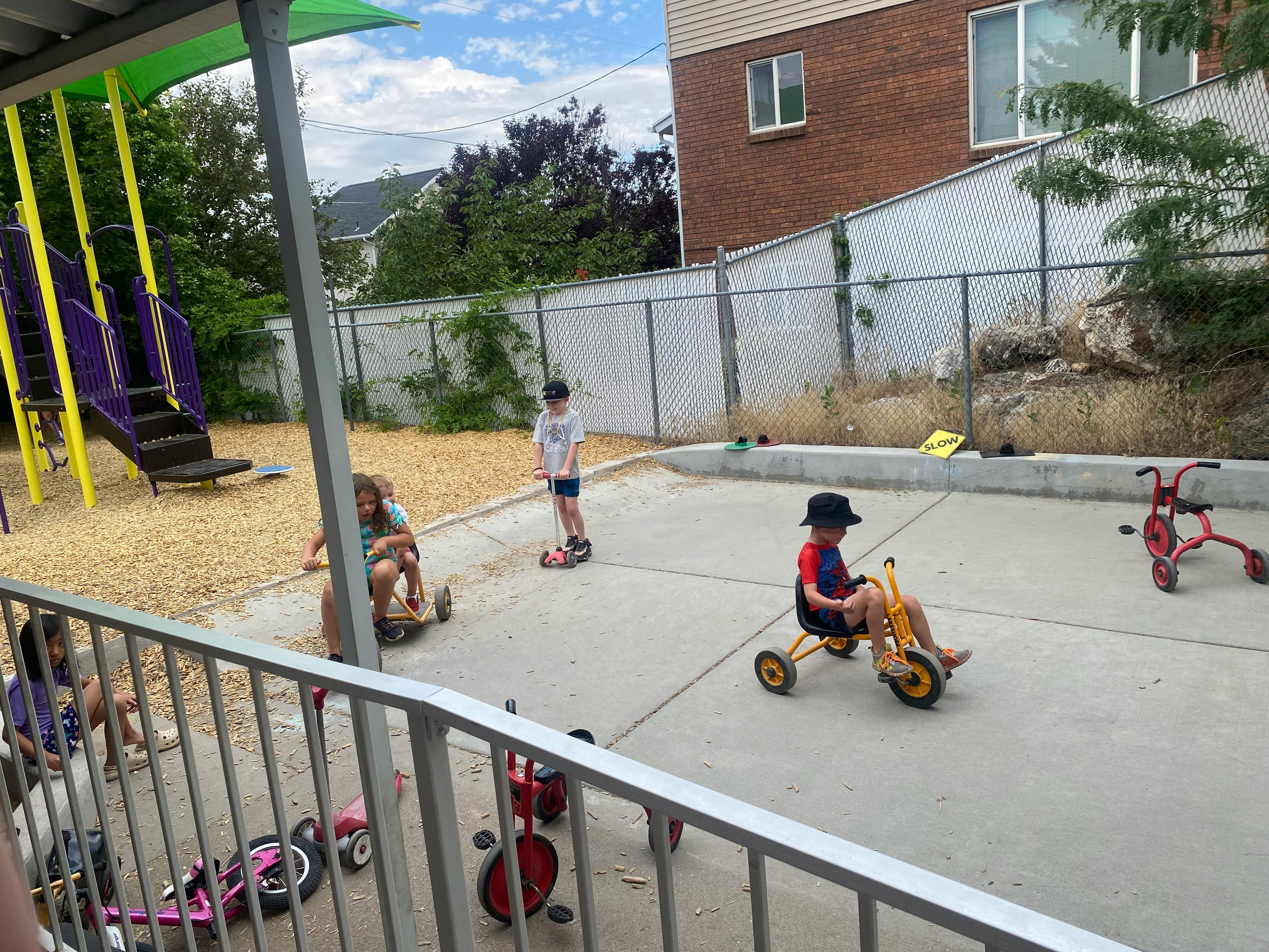 Children playing on scooters and tricycles on a paved area next to a playground